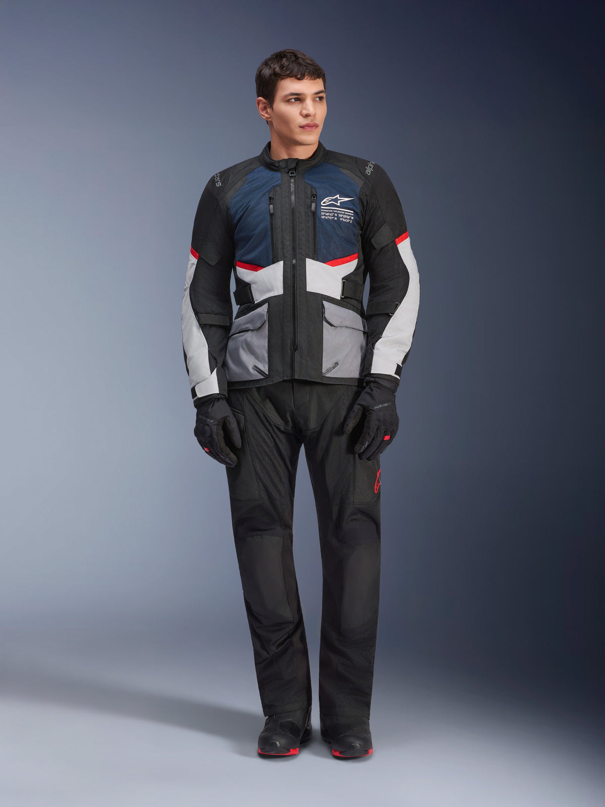 Veste Alpinestars Andes Air Drystar®, veste de touring moto, bleu foncé, noir et gris glace, portée avec un pantalon d'aventure et des gants noirs assortis, dotée de panneaux en mesh pour la circulation de l'air et un design technique multi-poches.