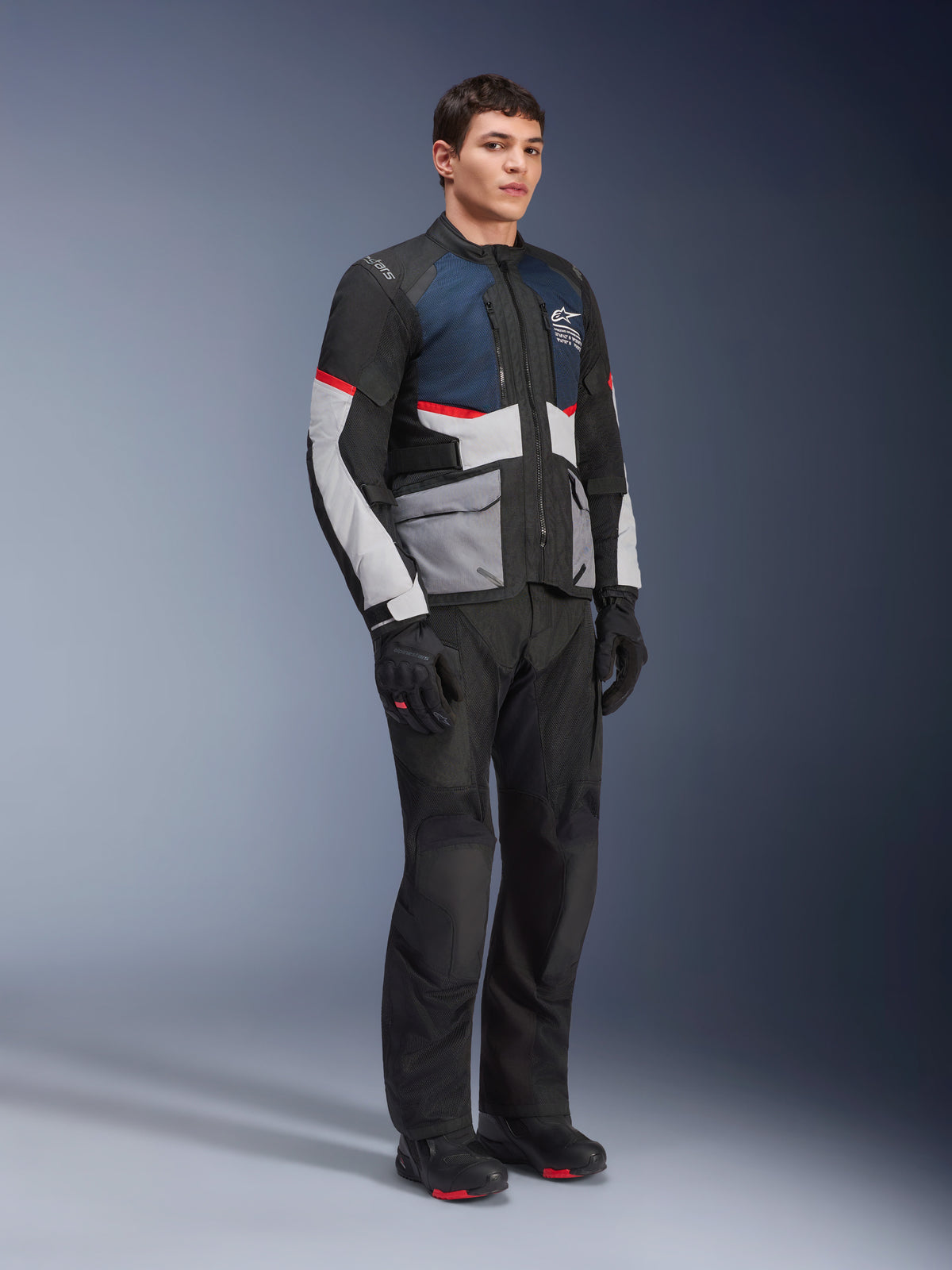 Modèle masculin portant la veste Alpinestars Andes Air Drystar® en Deep Blue Black Ice Gray, associée à un pantalon de moto, des gants et des bottes noirs, dotée de larges panneaux en maille pour la ventilation, de poches gris clair et de touches de rouge, conçue pour le touring d'aventure.