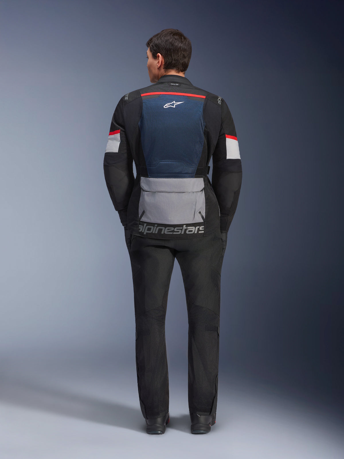 Veste Alpinestars Andes Air Drystar, Bleu Profond Noir Gris Glace, vue arrière sur mannequin, avec panneau arrière en maille bleue pour la ventilation, grande poche arrière grise, accents rouges et blancs sur les manches, logo « alpinestars », associée à un pantalon de moto noir pour une tenue de touring complète