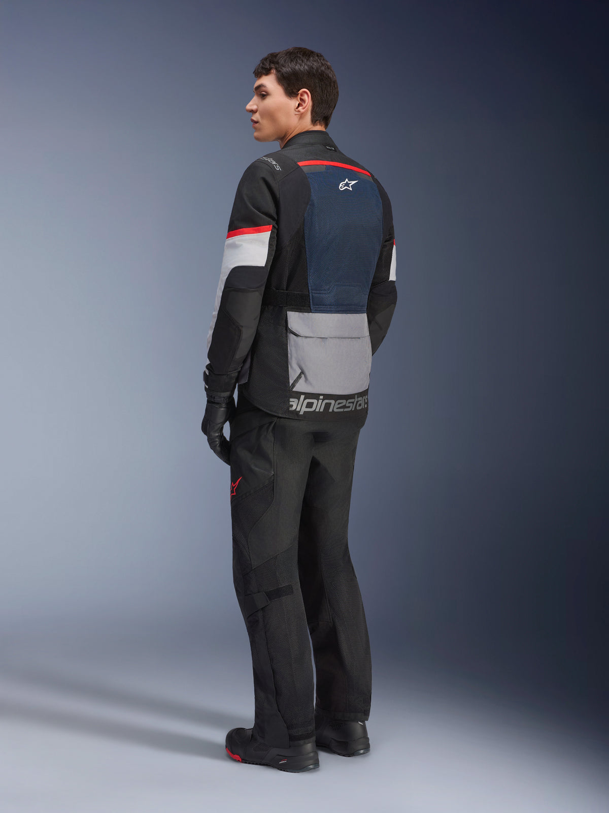 Modèle masculin vu de trois-quarts arrière portant la veste Alpinestars Andes Air Drystar® en Deep Blue Black Ice Gray, associée à un pantalon de moto et des gants noirs, avec empiècement dorsal en mesh bleu, poche utilitaire grise en bas du dos et accents rouges sur les manches, équipement de touring aventure professionnel.