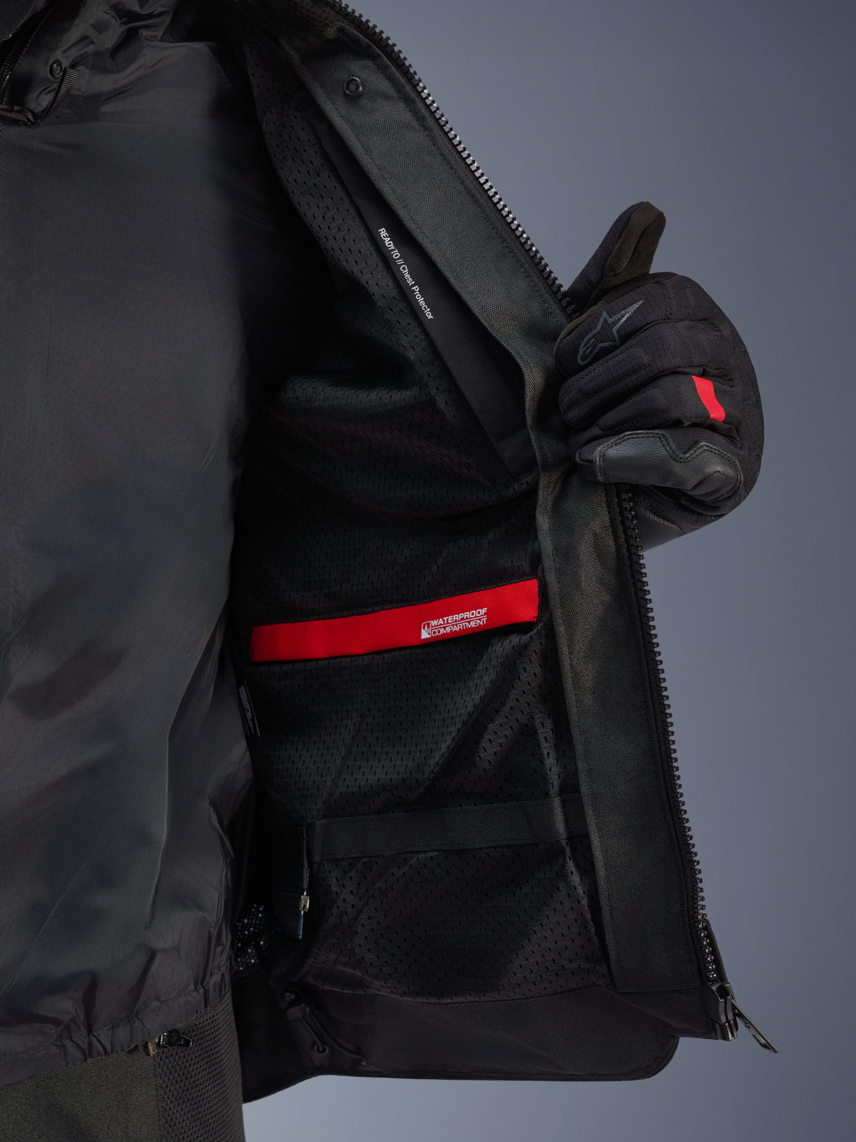 Vue détaillée de l'intérieur de la veste Alpinestars Andes Air Drystar®, mettant en évidence la doublure en maille noire respirante, le compartiment étanche rouge et la poche pour protection de poitrine, maintenue ouverte par un gant de moto noir.