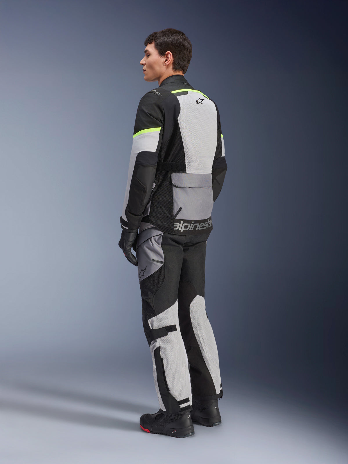 Motard portant une veste et un pantalon Alpinestars Andes Air Drystar®, équipement de touring aventure en Ice Gray Dark Gray Black avec des accents jaune fluo, doté de larges panneaux en mesh pour la circulation de l'air, logo Alpinestars au dos, porté avec des gants et des bottes noirs.