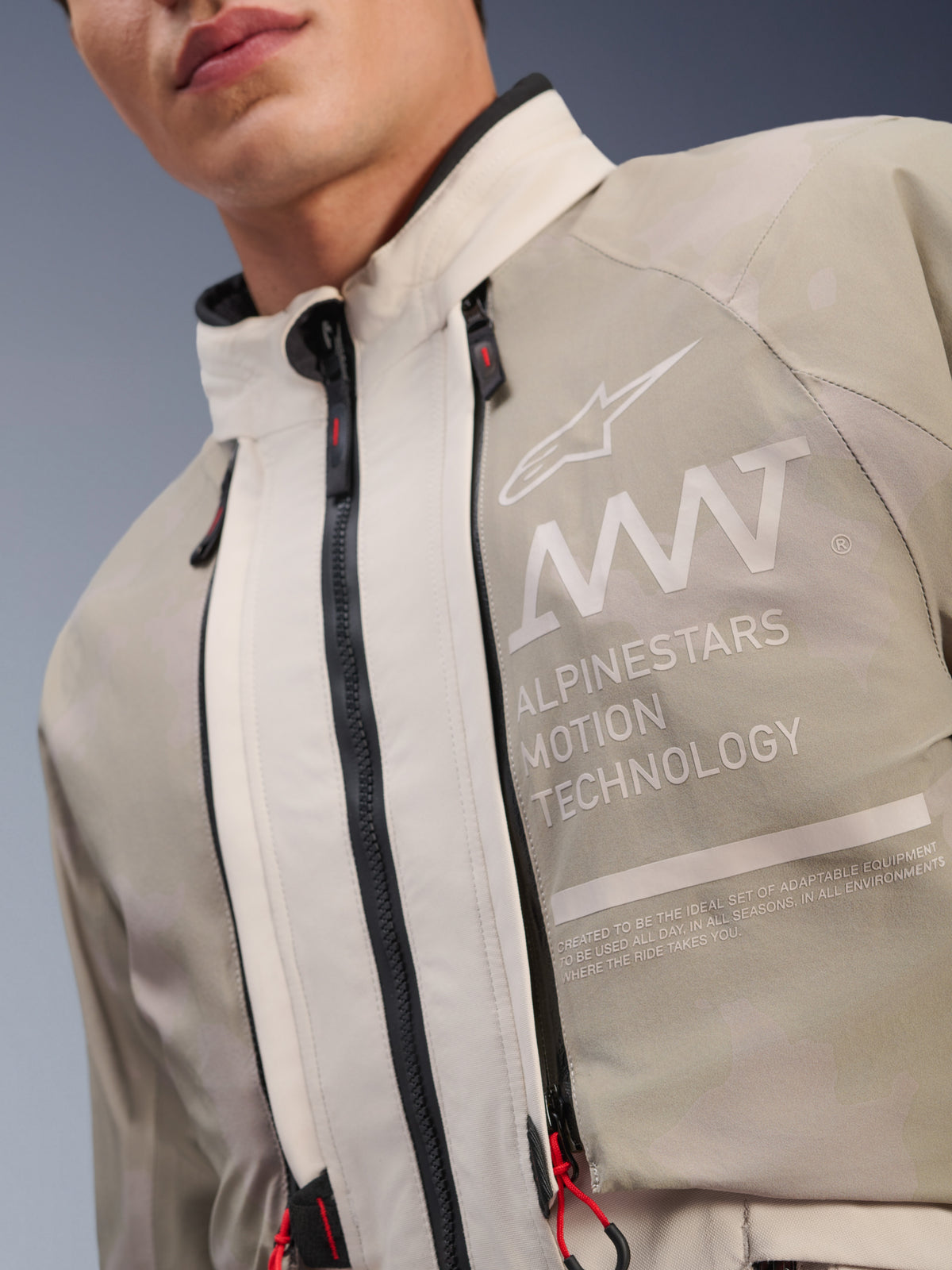Veste Alpinestars AMT-10LAB Drystar® XF, coloris White Sand Aluminum Camo, gros plan sur le torse avec marquage AMT Alpinestars Motion Technology, fermetures éclair imperméables à tirettes rouges et textile technique texturé.