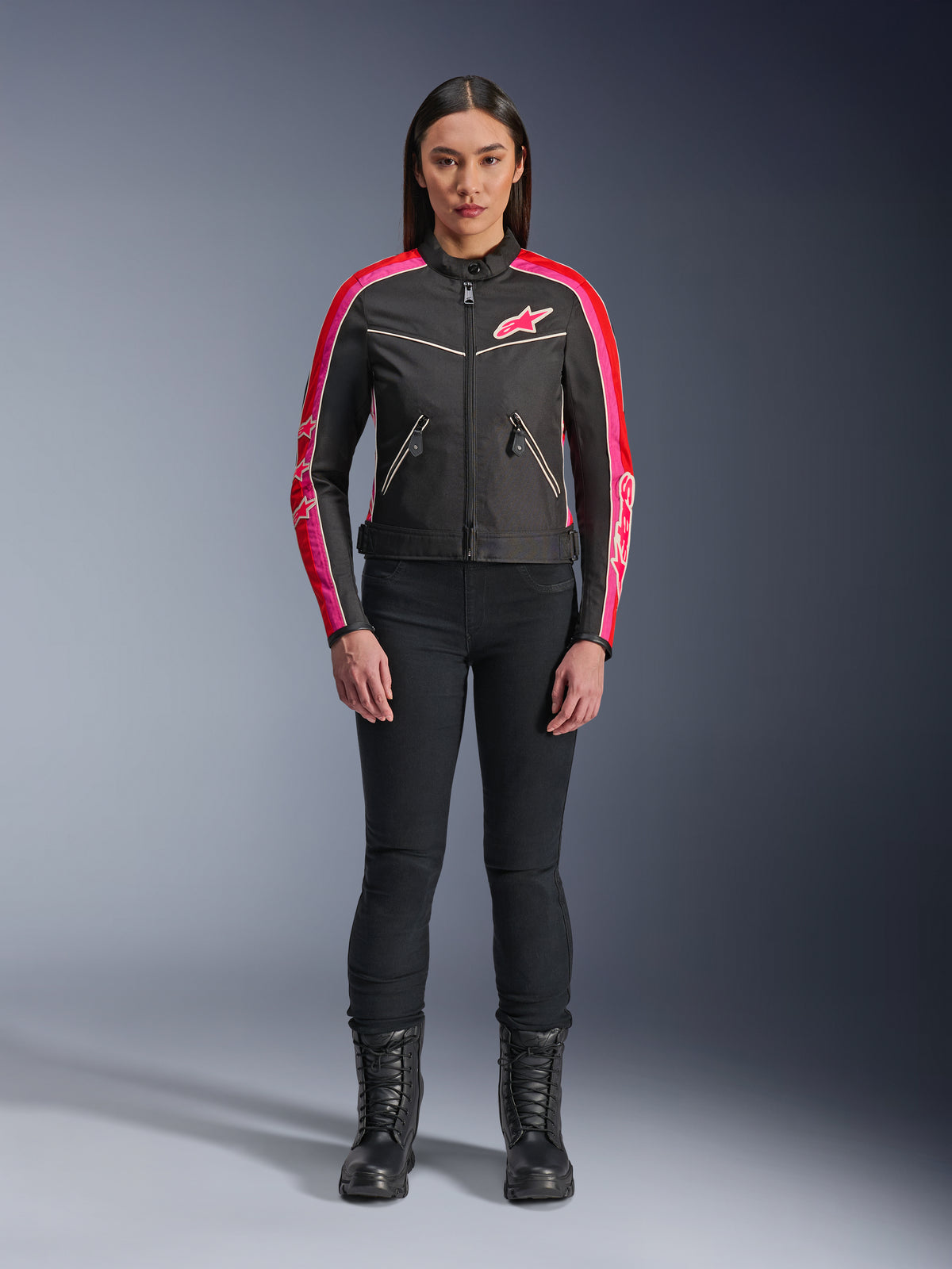 Femme portant la veste Alpinestars Stella T-Dyno WR, noire avec des accents rouge vif et fuchsia, veste textile de style vintage avec logo rose et bandes sur les manches, portée avec un pantalon noir et des bottes à lacets, équipement de moto urbain