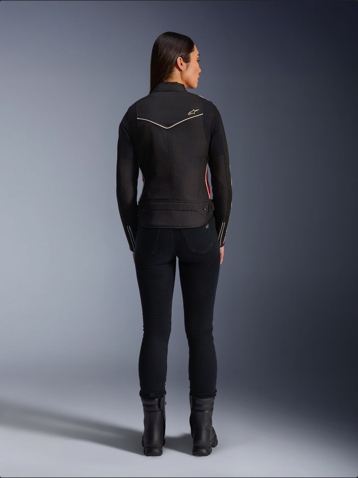 Modèle féminine de dos portant la veste Alpinestars Stella T-Dyno WR en Noir Rouge Éclatant Fuchsia, veste de moto en textile avec une coupe biker courte, vue arrière montrant le logo étoile blanc, associée à un pantalon noir et des bottes noires