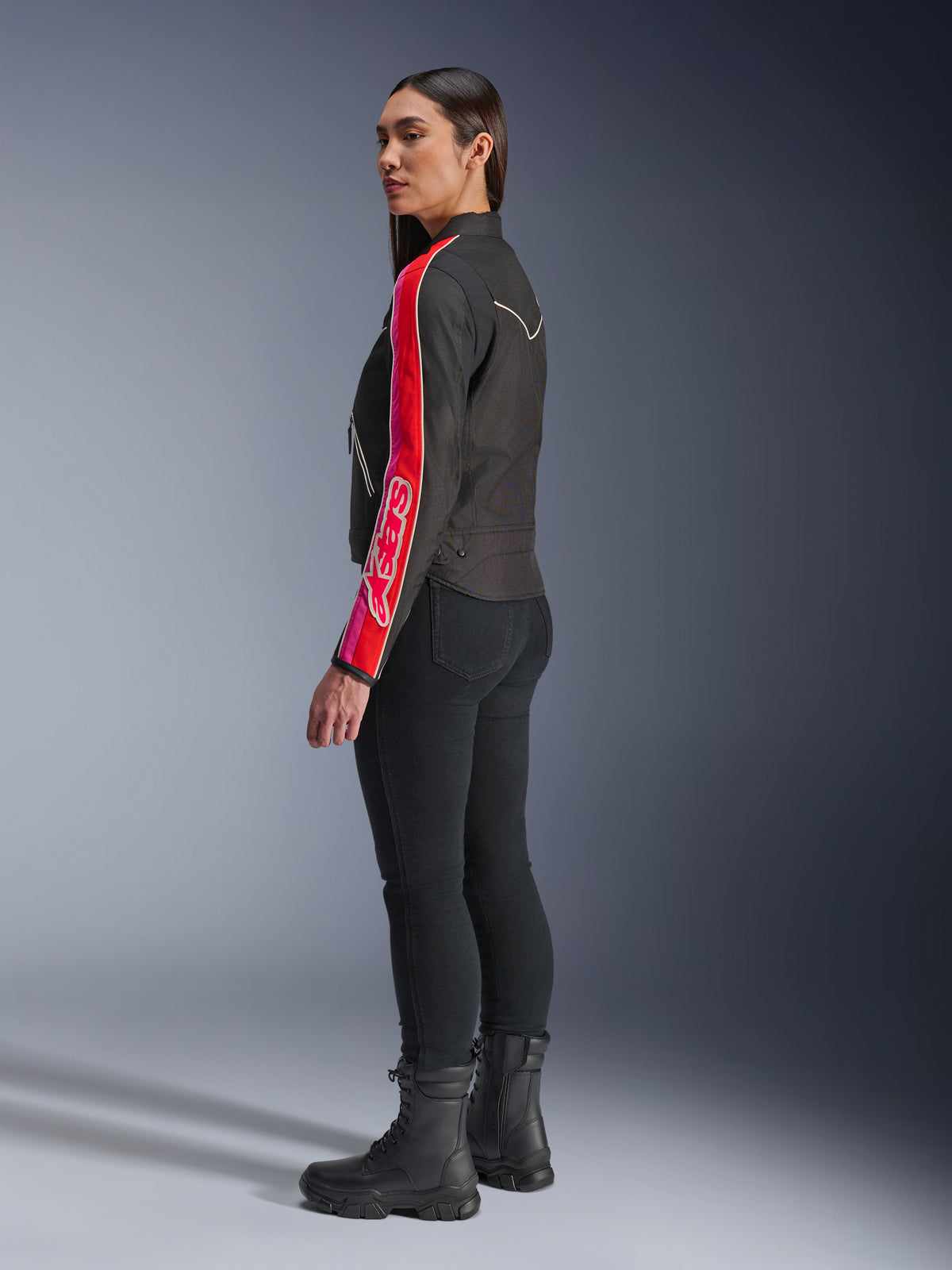 Modèle féminin portant la veste Alpinestars Stella T-Dyno WR en Noir Rouge Éclatant Fuchsia, avec des rayures rouges et roses vives et le logo Alpinestars sur la manche, associée à un pantalon de moto noir et des bottes de moto noires, en studio.