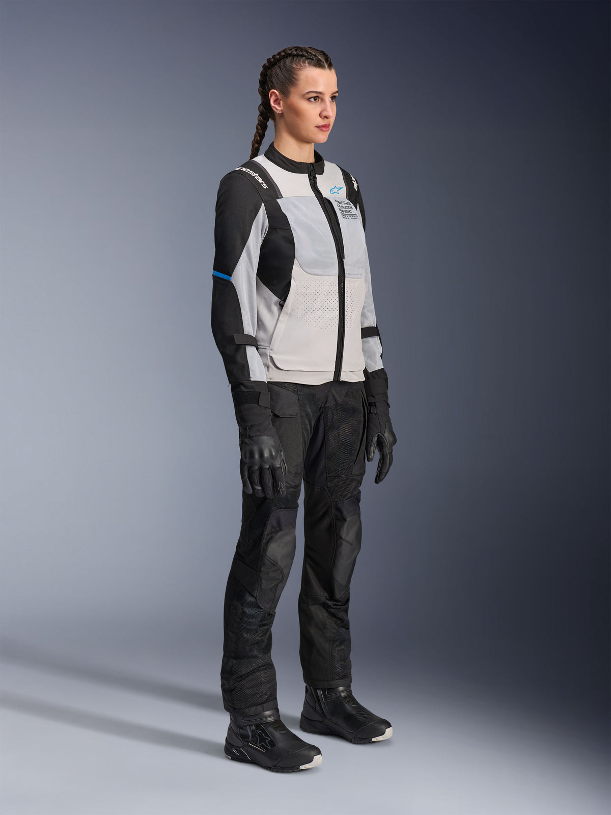 Mannequin portant la veste Alpinestars Stella ST-2 Air en Gris Glace et Noir, veste de moto technique pour femme avec panneaux en mesh et construction softshell, associée à un pantalon et des bottes de touring aventure noirs, présentant des accents bleus et un design de conduite moderne