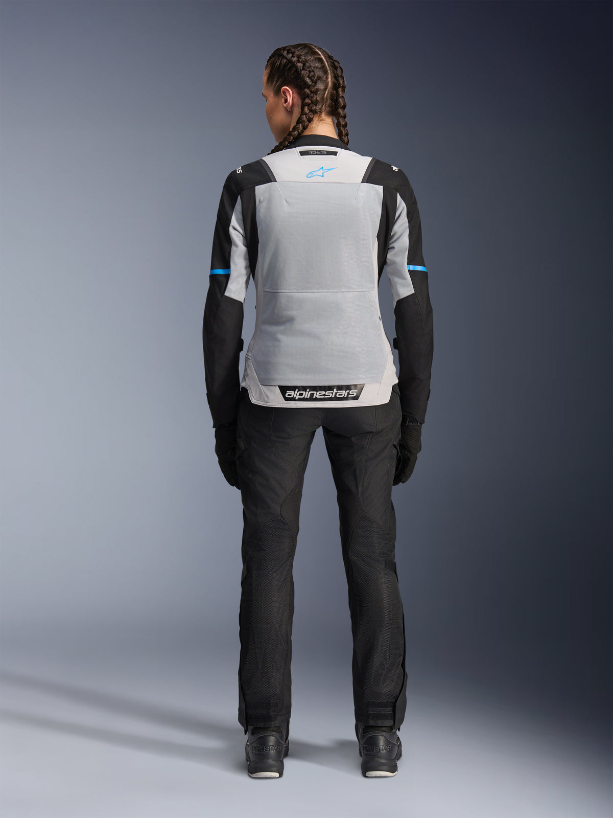 Veste Alpinestars Stella ST-2 Air, Ice Gray Black, vue arrière sur modèle féminin, avec un large panneau arrière en mesh gris clair, manches noires avec des bandes bleues, marquage Alpinestars sur le bas du dos, associée à un pantalon technique et des bottes de moto noirs, équipement de touring moto d'été