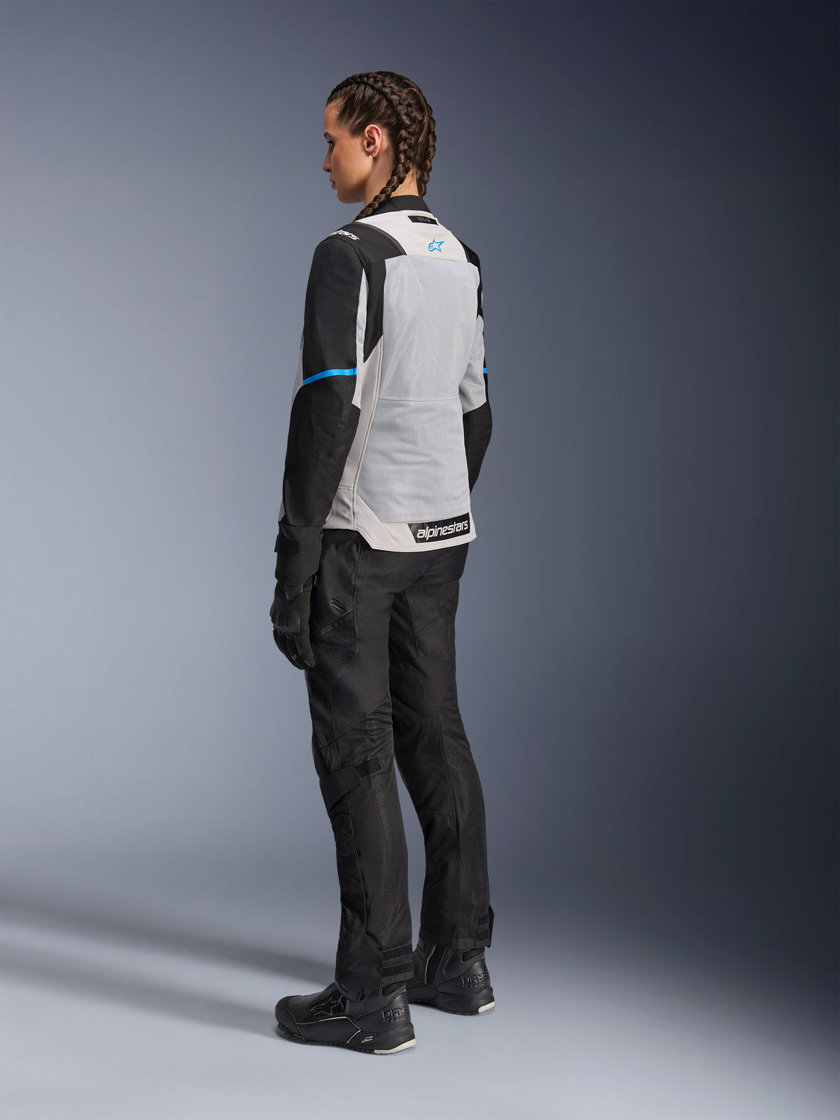 Motarde portant la veste Alpinestars Stella ST-2 Air en Gris Glace et Noir, vue de dos de trois-quarts mettant en valeur le large panneau arrière en maille gris clair, les manches noires avec une bande bleue et le logo Alpinestars bleu, associée à un pantalon, des gants et des bottes de moto noirs pour une tenue d'aventure touring complète.