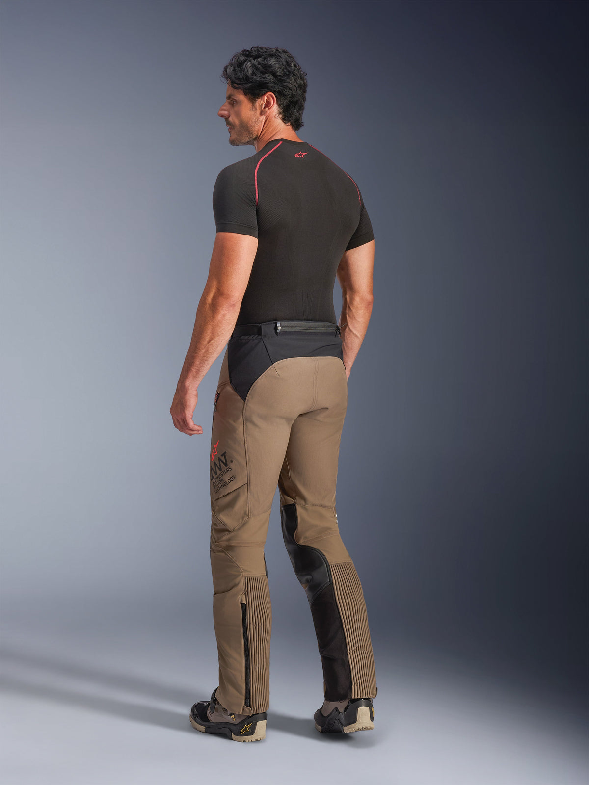 Pantalon Alpinestars AMT-8 Stretch Drystar® XF en Walnut Black, pantalon de moto adventure-touring vu de trois-quarts arrière, textile beige noyer avec empiècements noirs sur l'assise et l'intérieur des jambes, panneaux extensibles en accordéon aux mollets et logo AMT sur le côté, porté avec une couche de base noire.