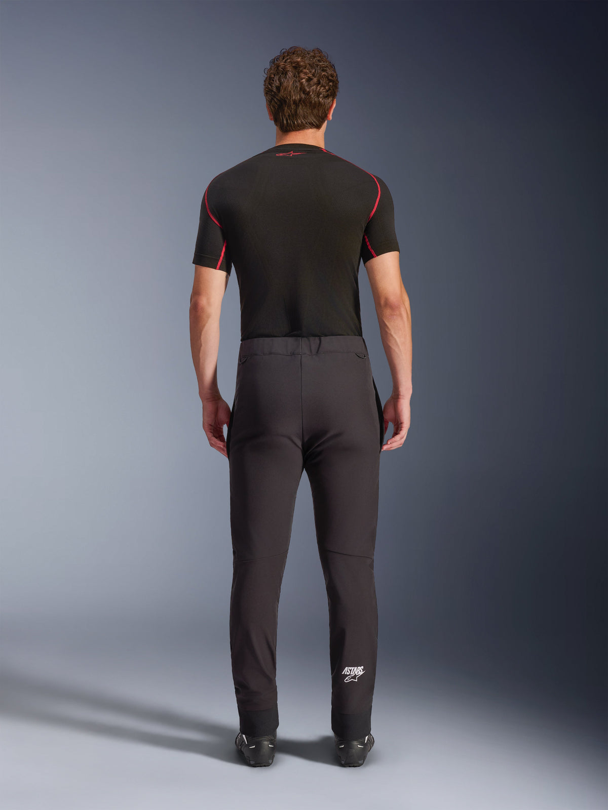 Pantalon Alpinestars Aeroshell, pantalon de moto softshell de sport polyvalent, noir, vue arrière, porté avec une couche de base noire à manches courtes, avec une coupe ajustée, des genoux pré-courbés et un logo Astars blanc sur le bas de la jambe gauche.