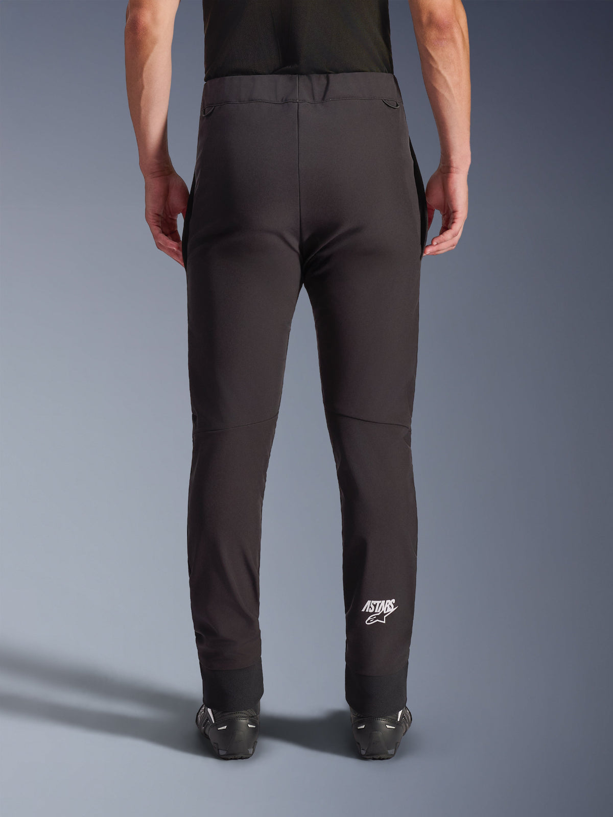 Pantalon Alpinestars Aeroshell, pantalon de moto en softshell noir, vue arrière montrant la coupe ajustée et les genoux pré-courbés, logo Astars blanc sur le bas du mollet droit, ourlet inférieur élastique, design polyvalent pour la route