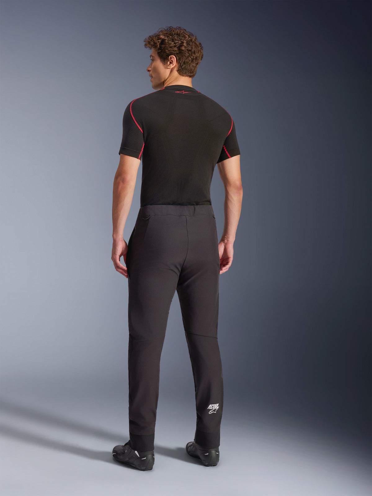 Pantalon Aeroshell Alpinestars, noir, pantalon de moto softshell, vue de dos sur un mannequin, doté d'une coupe fuselée moderne, d'un logo Astars blanc sur le bas de la jambe et d'ourlets élastiqués, porté avec une couche de base noire à manches courtes et des chaussures de moto