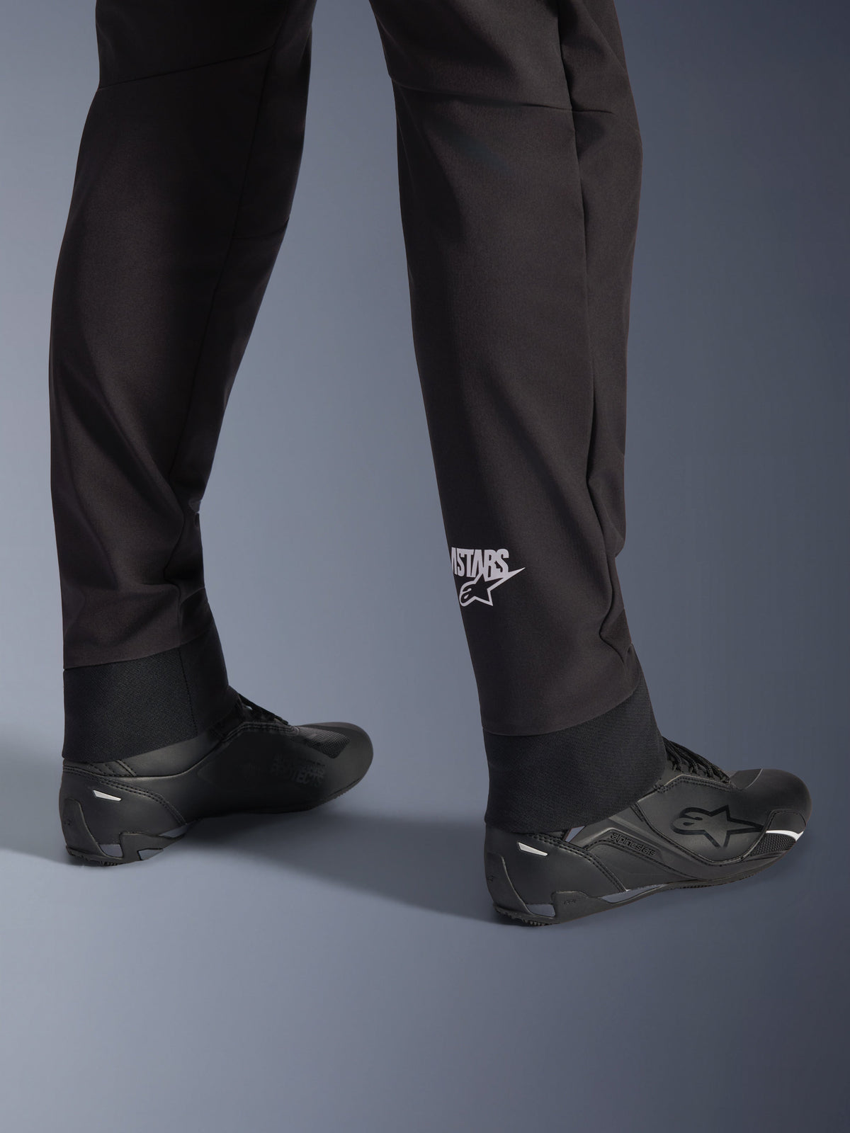 Pantalon Alpinestars Aeroshell, pantalon de moto softshell noir, gros plan sur le bas de la jambe et l'ourlet élastiqué, logo Alpinestars blanc sur le mollet, porté avec des chaussures de moto noires, design crossover sport moderne