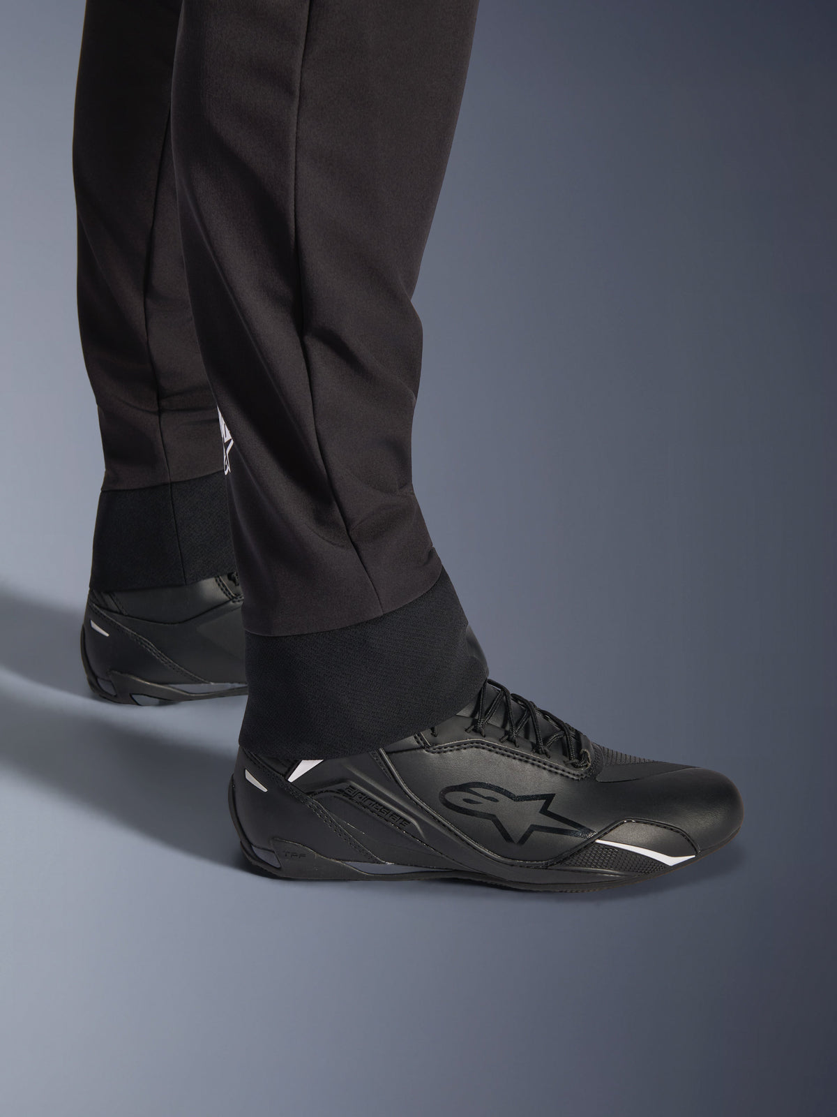 Pantalon Alpinestars Aeroshell, noir, vue rapprochée de l'ourlet inférieur et du revers élastiqués, design softshell sportif moderne, porté avec des chaussures de moto Alpinestars noires