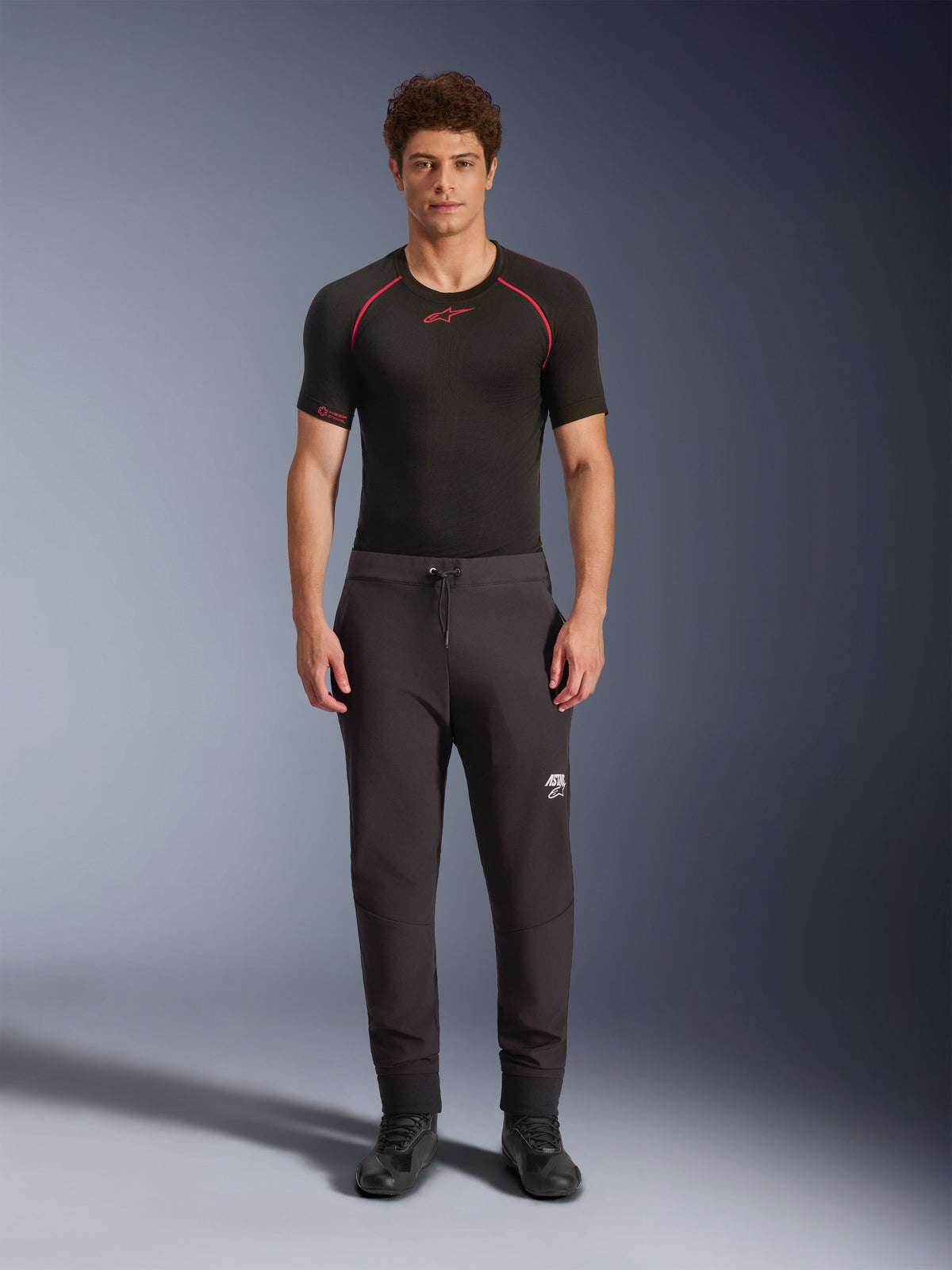 Pantalon Alpinestars Aeroshell, pantalon de moto softshell noir, porté avec une couche de base noire à manches courtes et des chaussures noires, avec taille élastique à cordon de serrage, logo Astars blanc sur la cuisse et ourlets élastiques, silhouette athlétique moderne pour la route