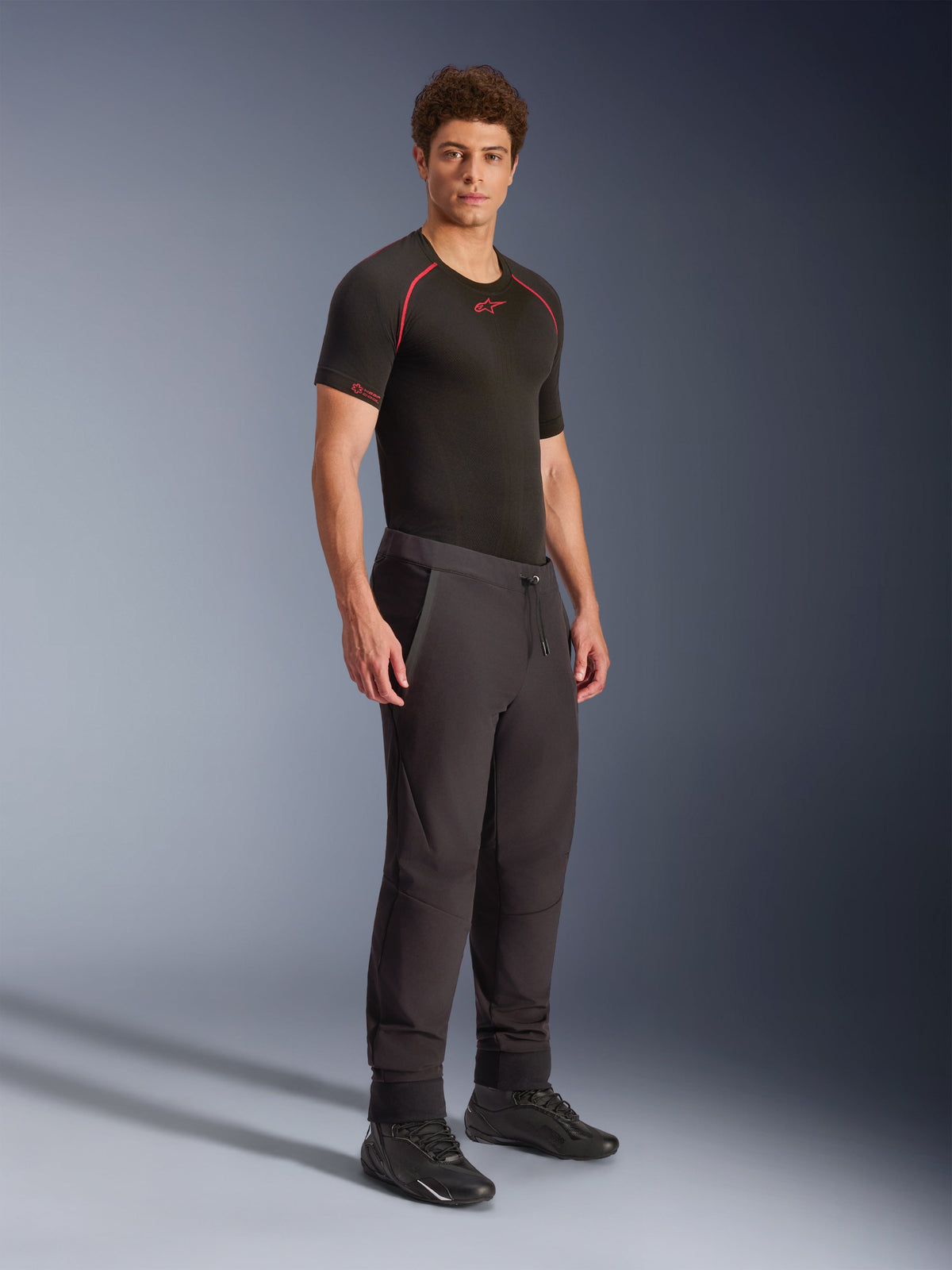 Pantalon Alpinestars Aeroshell, noir, pantalon de moto softshell sport polyvalent, porté par un mannequin homme avec une couche de base technique noire et des chaussures noires, doté d'une taille avec cordon de serrage et d'ourlets élastiques.