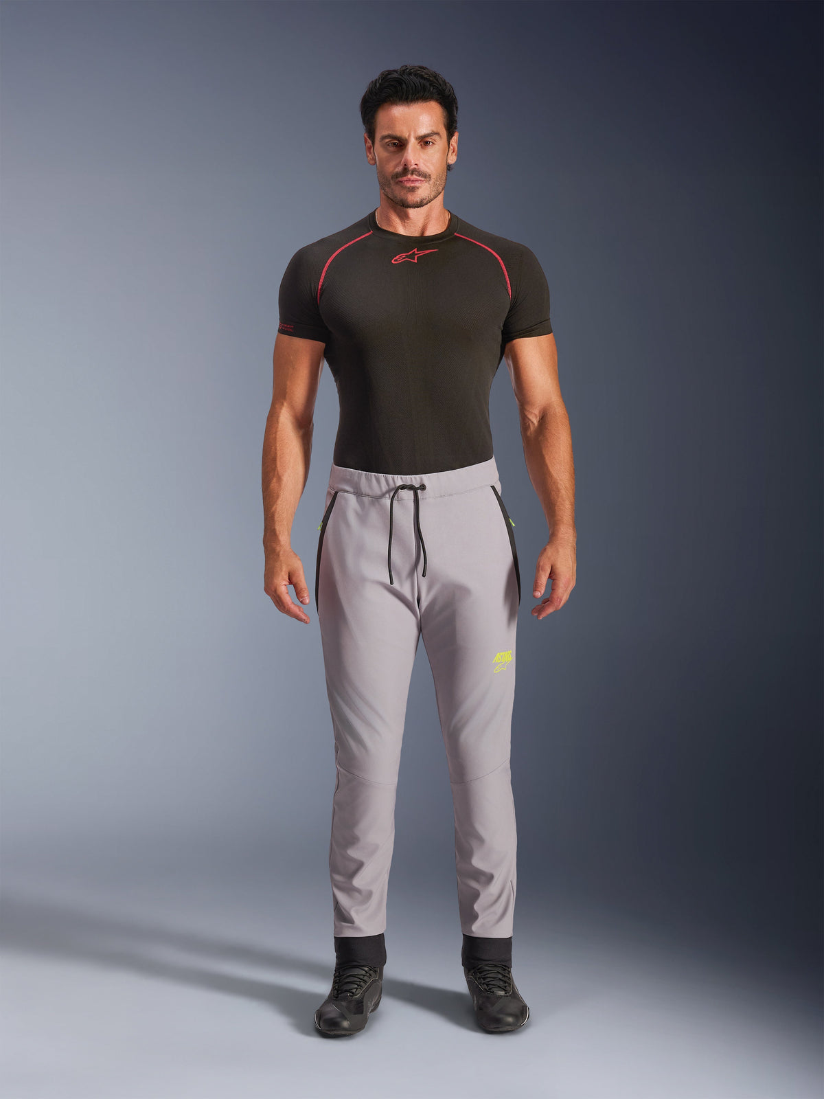 Mannequin homme portant le pantalon Aeroshell Alpinestars en Gris Moyen et Jaune Fluo, pantalon softshell de sport polyvalent avec taille élastique à cordon de serrage et poches zippées, associé à un haut technique noir à manches courtes, design athlétique moderne pour la route et la conduite décontractée