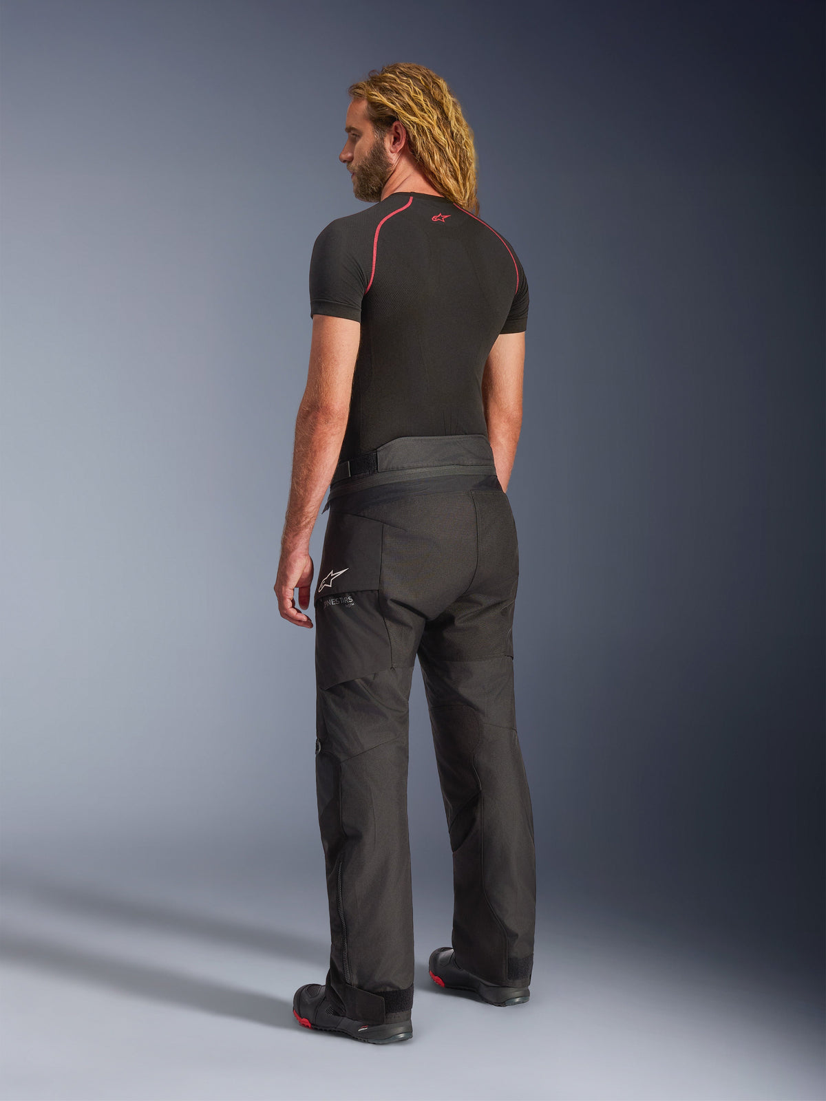 Pantalon Alpinestars Andes V4 Drystar® - Long, pantalon de moto d'aventure, noir, vue de trois-quarts arrière sur mannequin, design textile durable avec panneaux renforcés à l'assise et aux genoux, logo étoile Alpinestars, porté avec une sous-couche noire et des bottes de moto