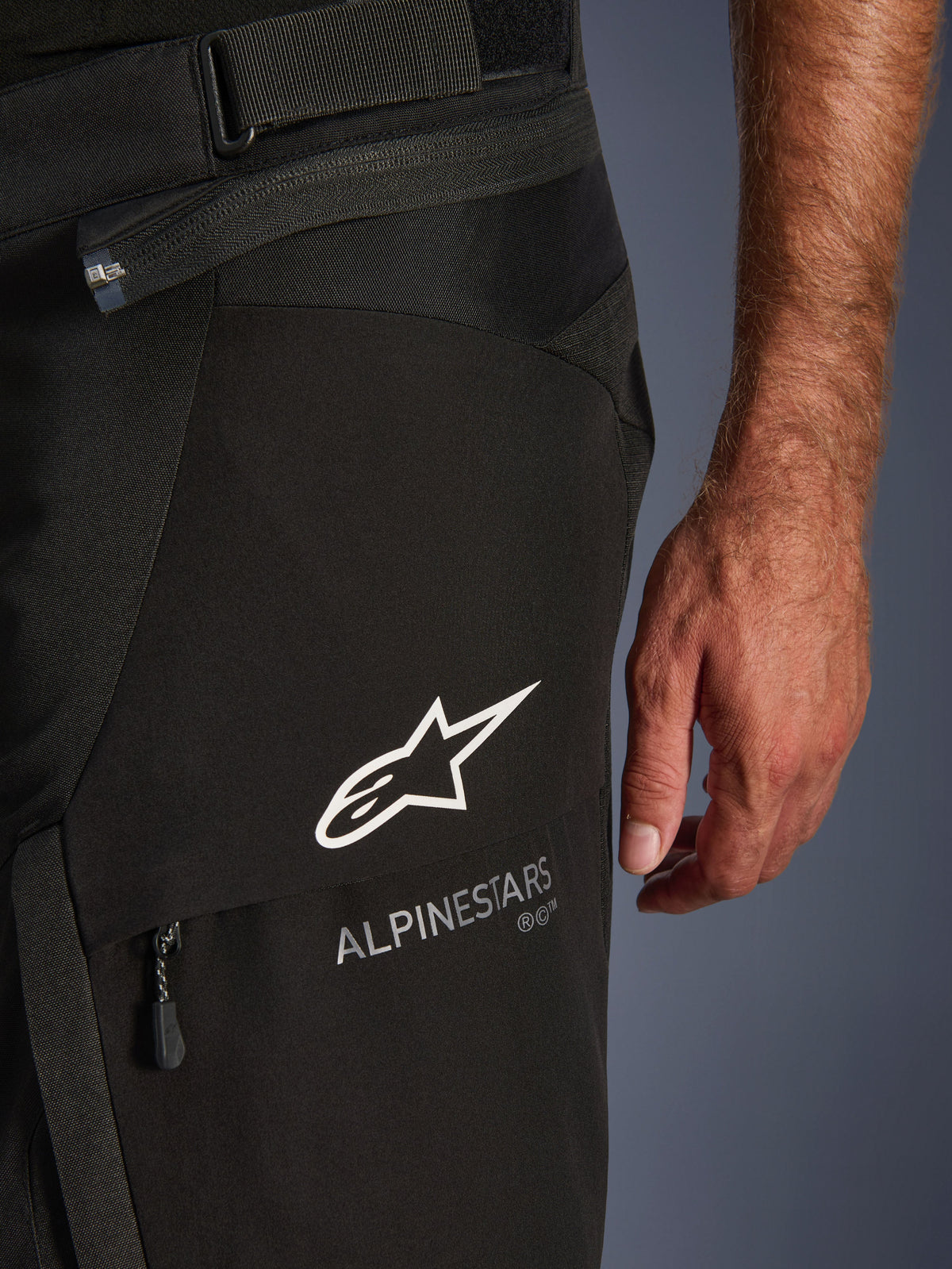 Pantalon Alpinestars Andes V4 Drystar, pantalon d'aventure moto, noir, gros plan sur le haut de la cuisse montrant le logo étoile et le texte Alpinestars blancs, sangle de réglage à la taille et détail de la poche zippée.