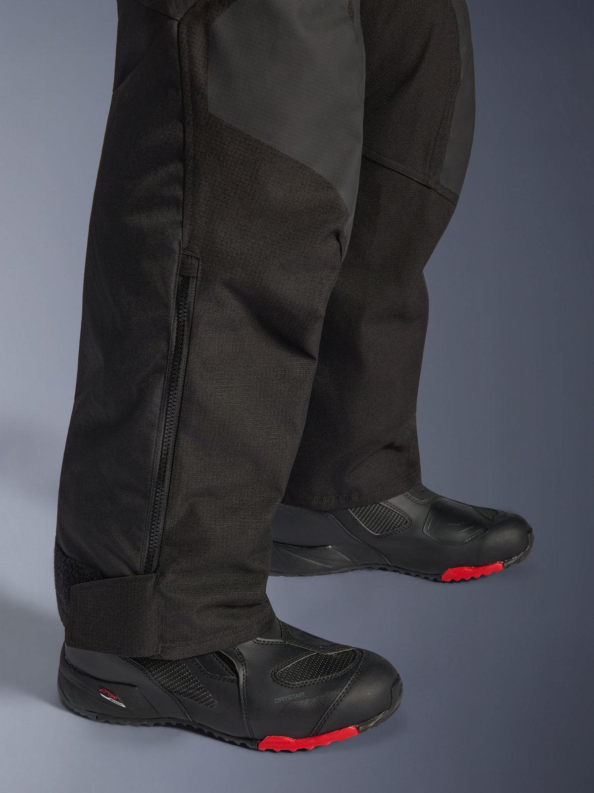 Pantalon Alpinestars Andes V4 Drystar® - Long, pantalon de moto d'aventure noir, gros plan sur le bas de la jambe montrant le revers zippé ajustable et la construction textile durable, porté avec des bottes de moto noires avec des accents rouges