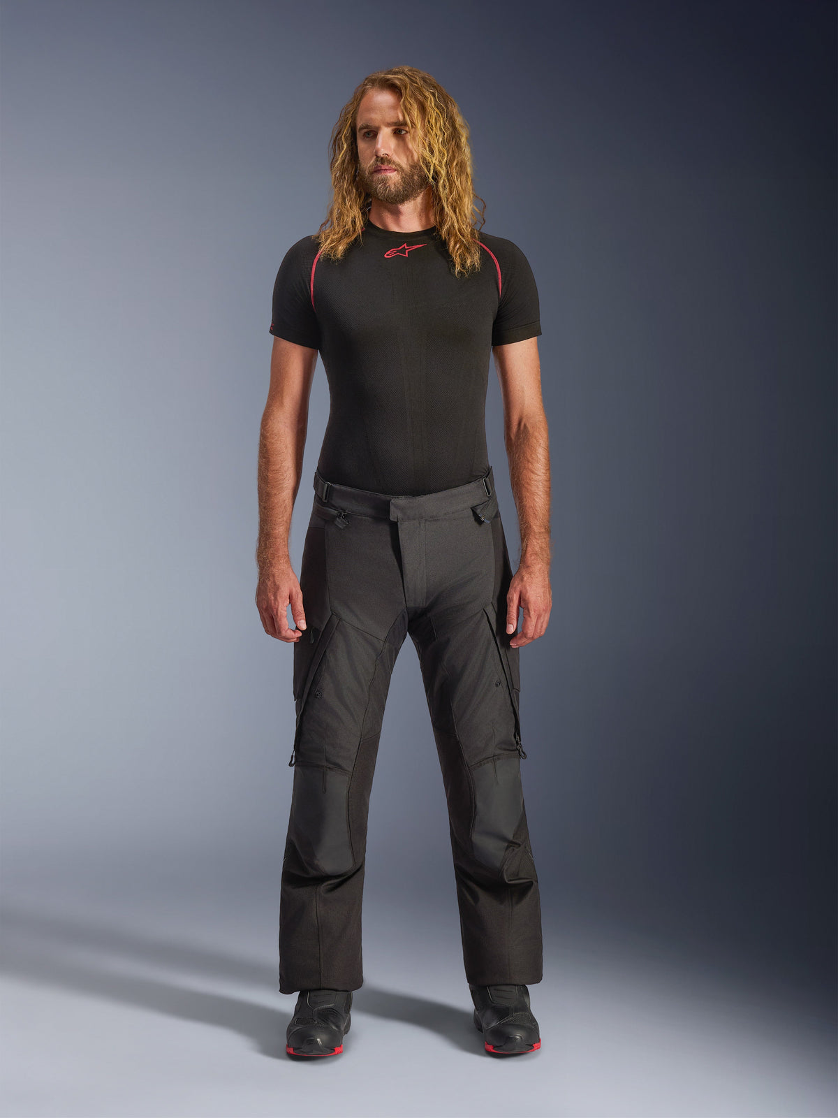 Modèle masculin portant le pantalon Alpinestars Andes V4 Drystar® - Long en noir, un pantalon de moto adventure-touring doté de panneaux de genoux renforcés et de poches cargo, associé à une couche de base noire à manches courtes et des bottes de moto noires.