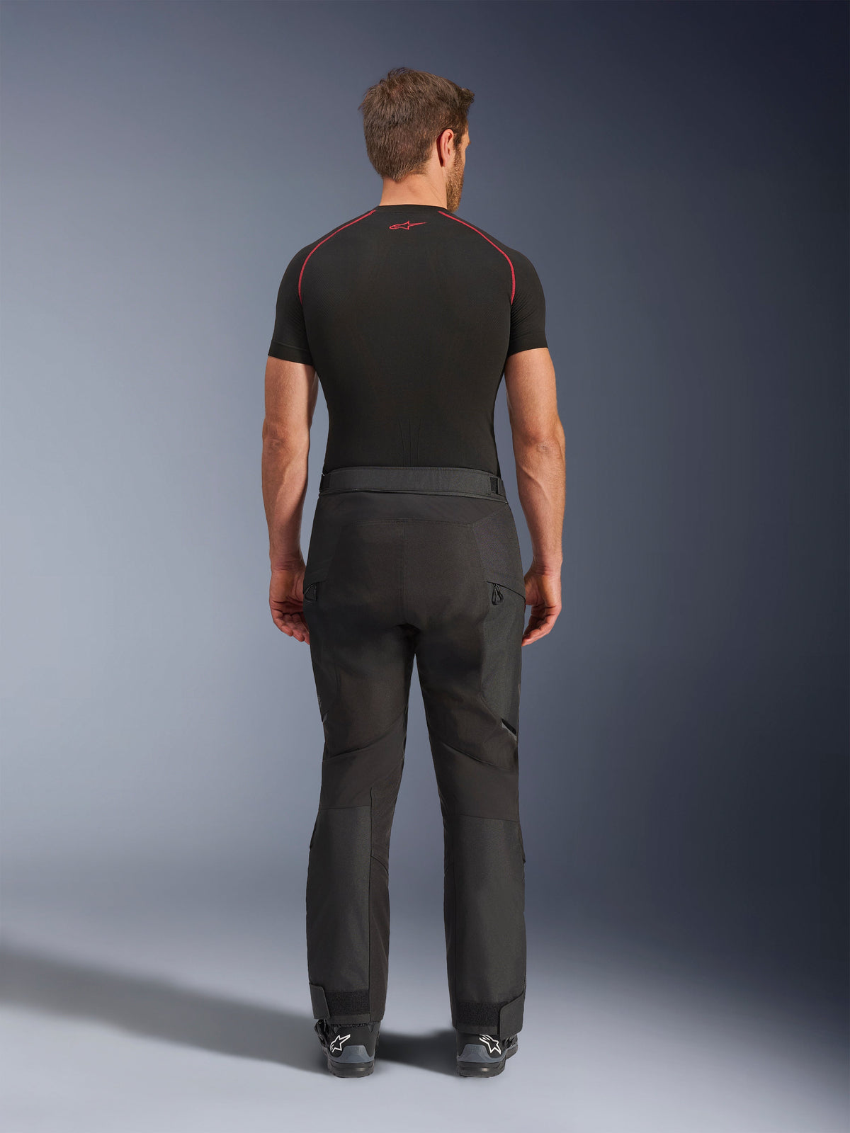 Rider wearing Alpinestars Cusco Drystar® Pants, Black adventure motorcycle pants, back view, paired with a black short-sleeve compression shirt and black riding boots, featuring a durable textile design with articulated panels for touring
