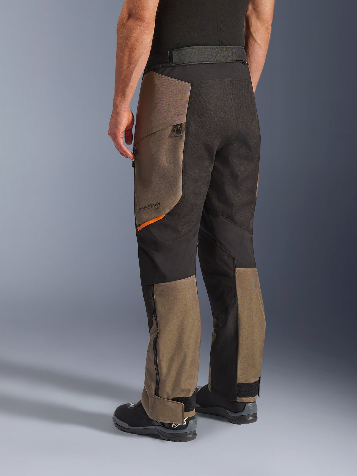 Pantalon Alpinestars Cusco Drystar®, pantalon de moto d'aventure, Walnut Black avec petit accent orange, vue de trois quarts arrière montrant la construction textile et les panneaux noyer, porté avec des bottes de moto noires