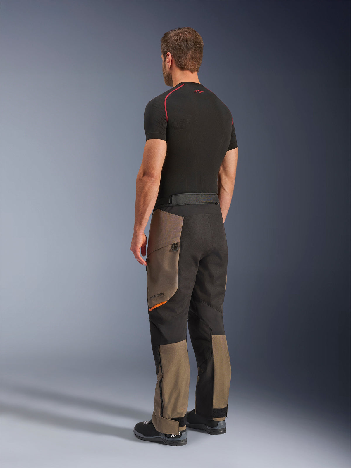 Pantalon Alpinestars Cusco Drystar®, pantalon de moto aventure, Walnut Black, vue de trois-quarts arrière porté avec une couche de base noire et des bottes, doté de panneaux textiles renforcés, de poches cargo zippées sur les cuisses et d'une coupe touring ergonomique.