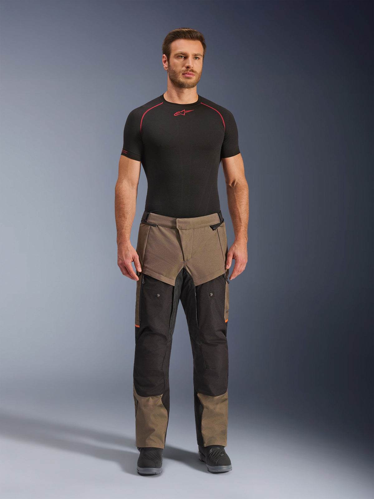 Pantalon Alpinestars Cusco Drystar®, Walnut Black, pantalon de moto adventure touring, porté par un mannequin homme avec une couche de base noire et des bottes noires, avec des panneaux noyer et noirs, des accents orange et des fermetures éclair de ventilation directe sur les cuisses.
