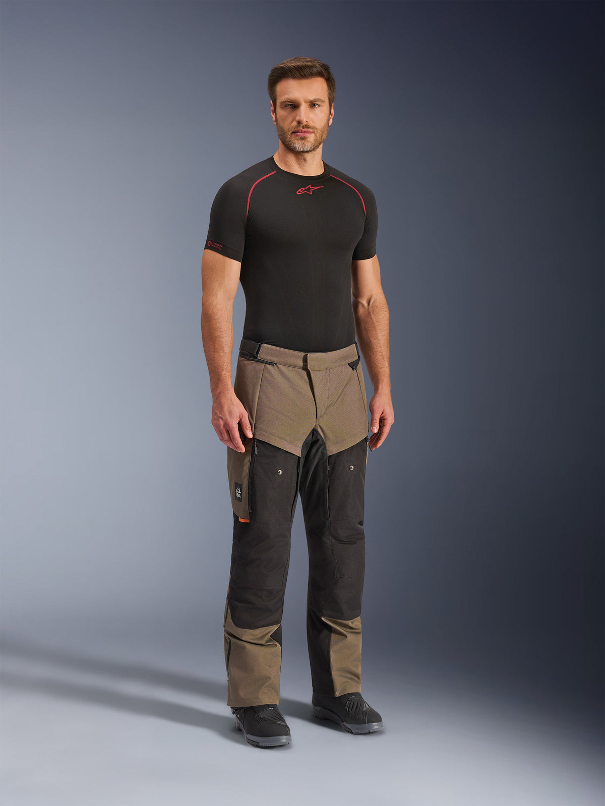 Pantalon Alpinestars Cusco Drystar®, pantalon de moto aventure, noyer et noir, porté par un mannequin avec une couche de base noire et des bottes, doté de grandes poches cargo sur les cuisses, de fermetures éclair de ventilation et d'un design bicolore robuste.