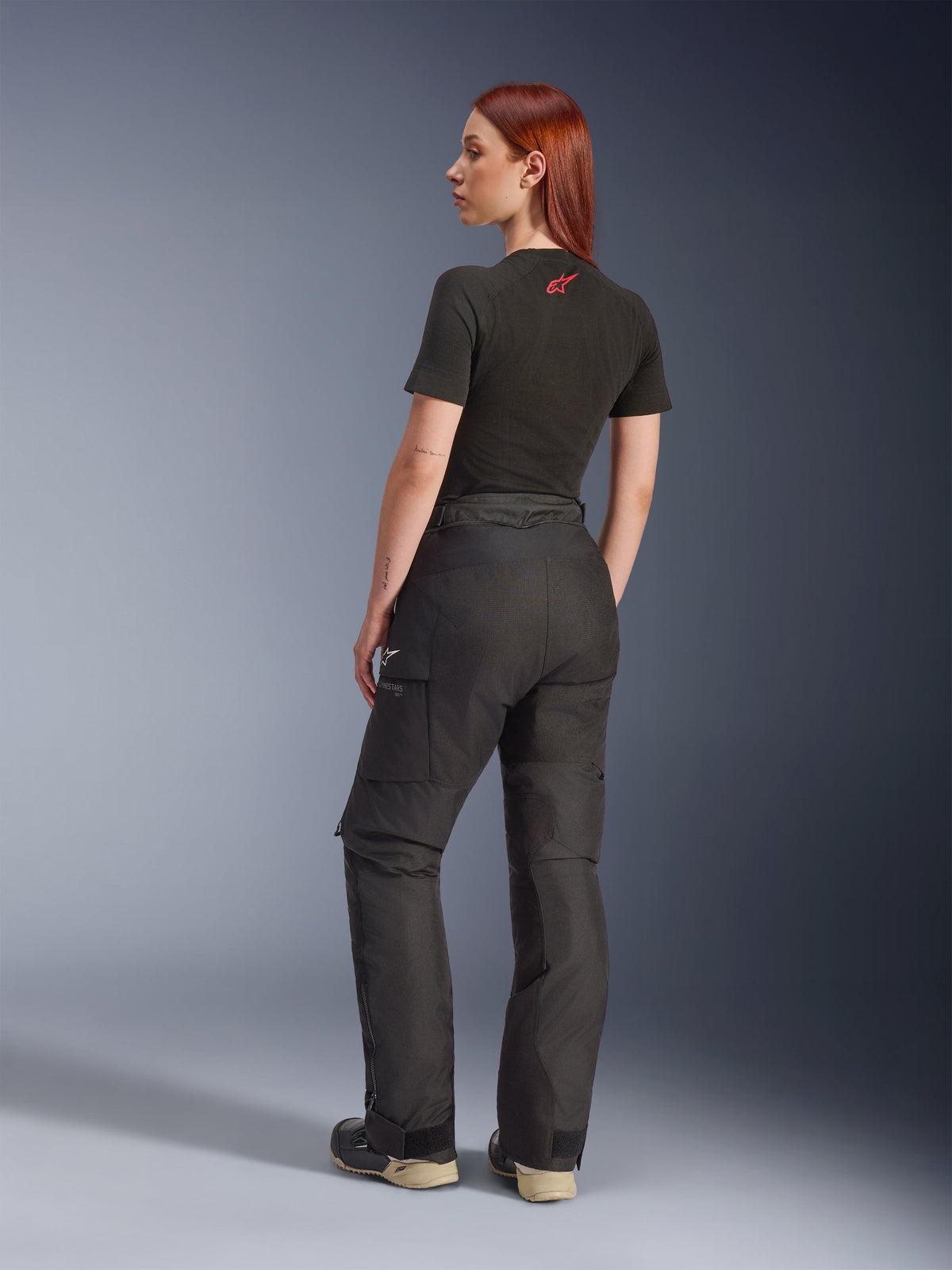 Pantalon Alpinestars Stella Andes V4 Drystar®, noir, pantalon de moto aventure présenté de trois-quarts arrière sur un mannequin, associé à un t-shirt noir et des bottes, doté de poches cargo latérales avec logo blanc et une construction en textile durable.