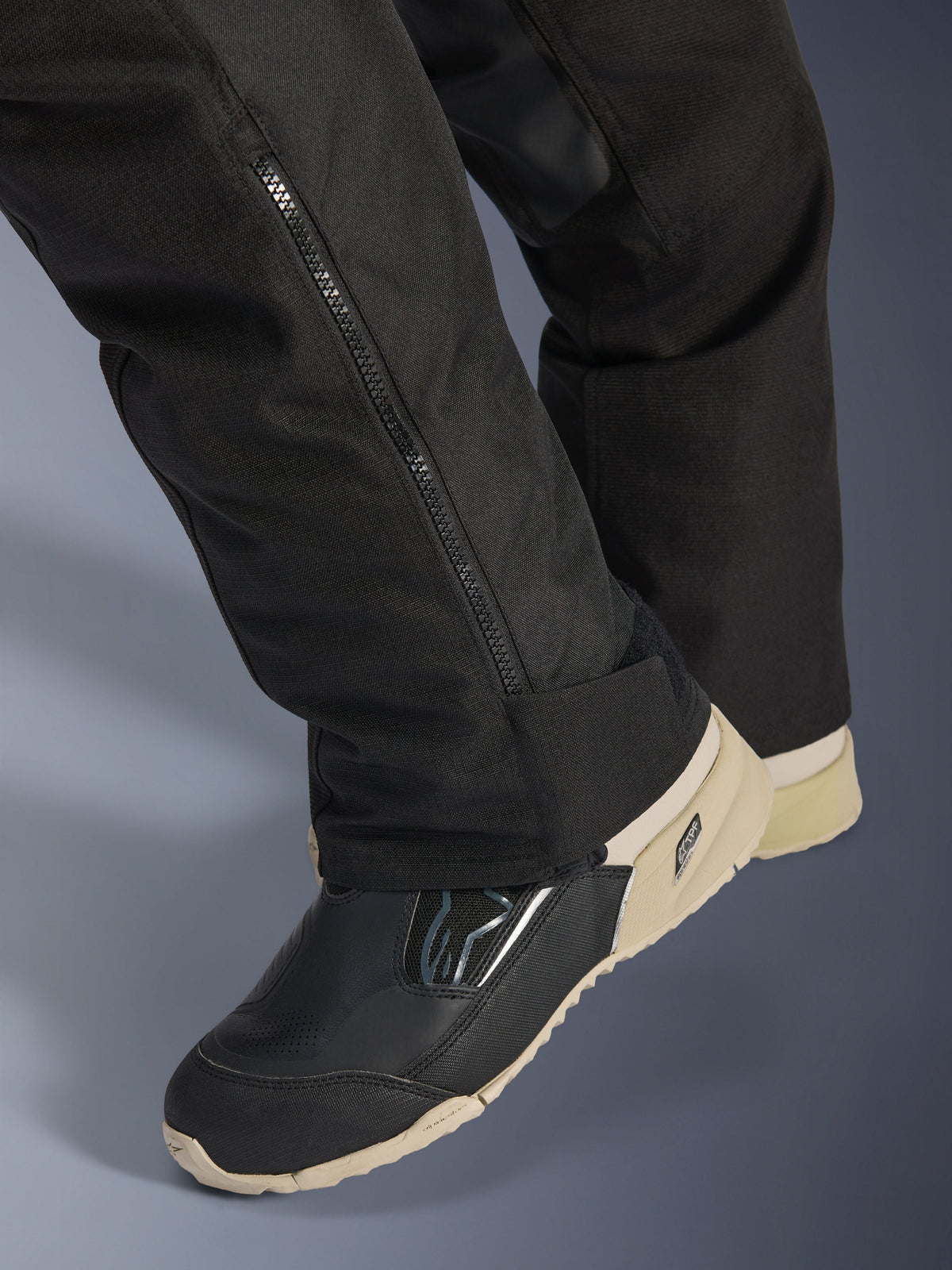 Pantalon Alpinestars Stella Andes V4 Drystar®, noir, gros plan sur le bas de la jambe avec fermeture éclair latérale pour l'ajustement des bottes, matériau textile durable, porté avec des bottes de moto noires et sable, design fonctionnel prêt pour l'aventure