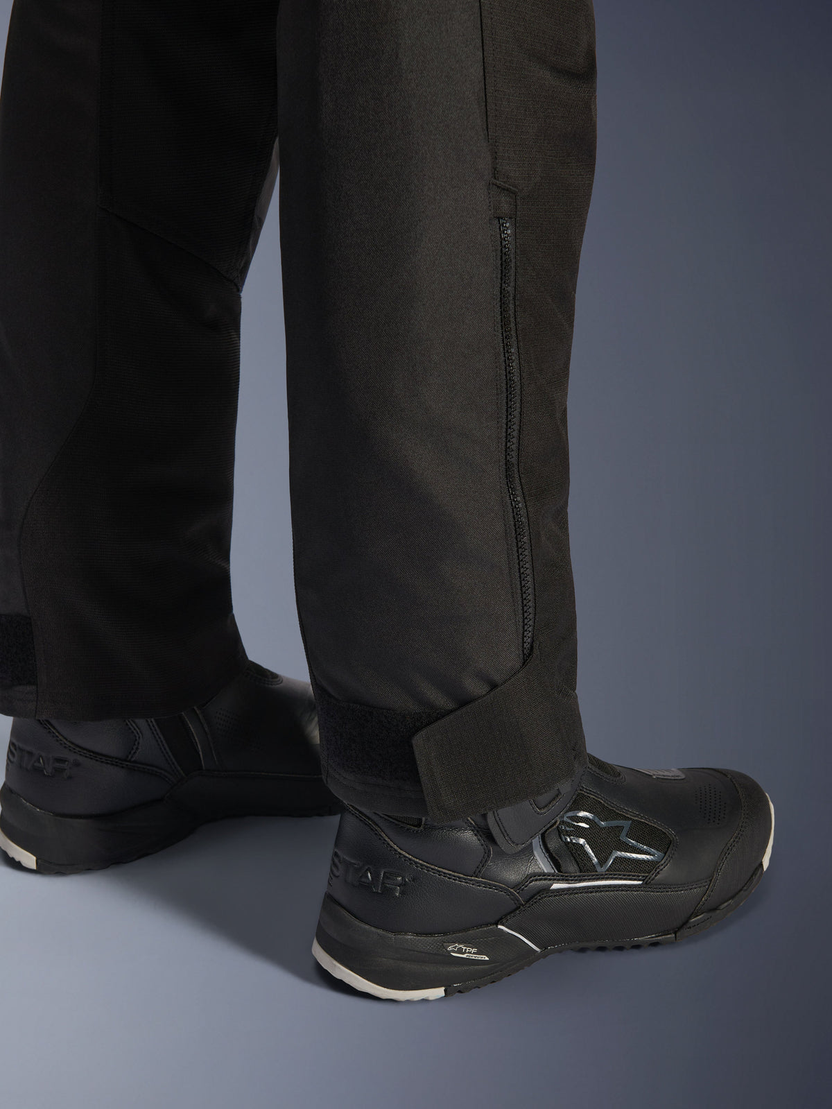 Pantalon Alpinestars Stella Andes V4 Drystar®, noir, vue rapprochée du bas de la jambe et de la cheville, avec fermeture éclair latérale et sangle de cheville réglable auto-agrippante, porté avec des bottes de moto Alpinestars noires