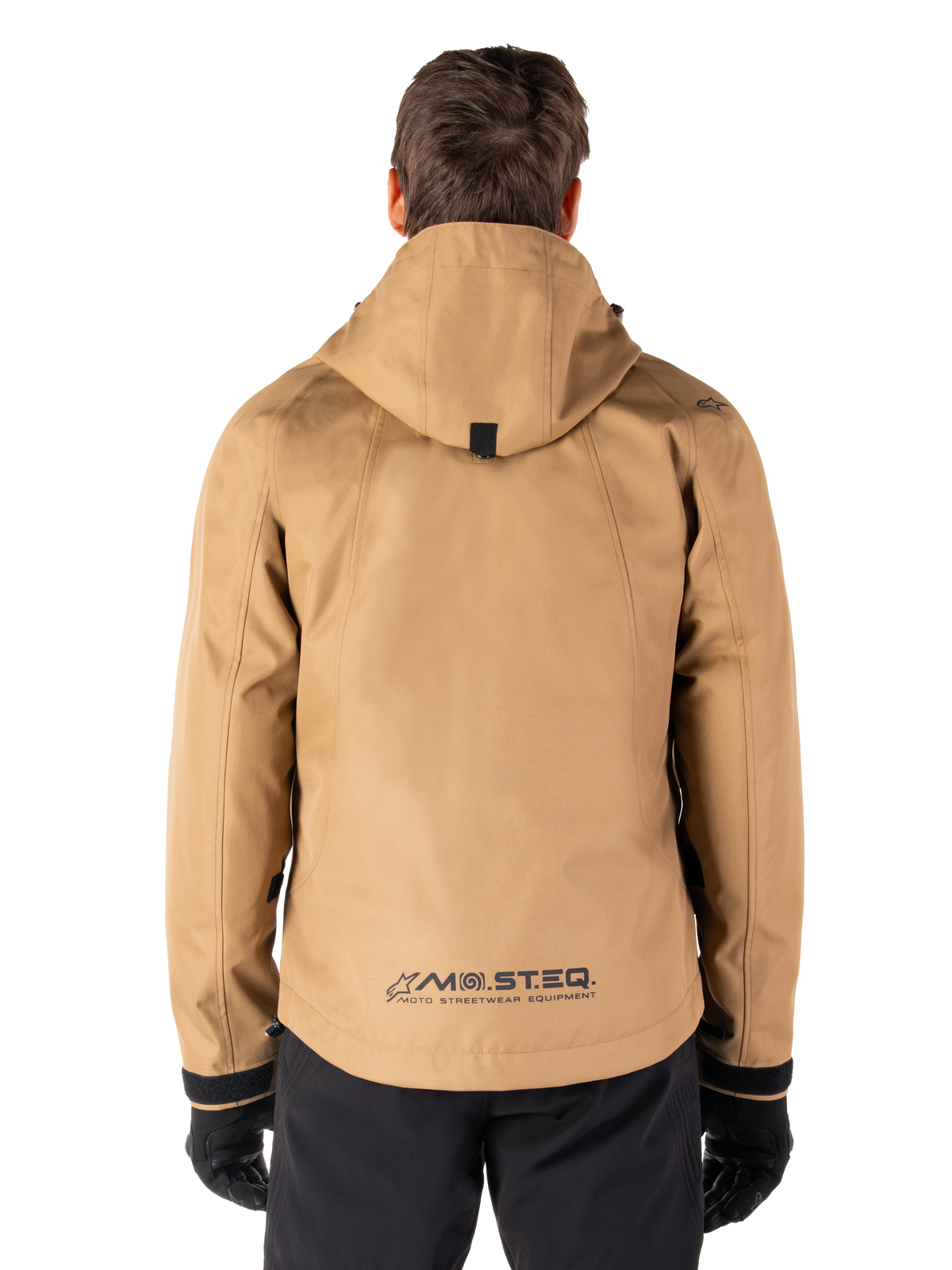 Parka imperméable Alpinestars Morush, Brun, vue arrière, dotée d'une capuche et du marquage MO.ST.EQ. en bas du dos, associée à un pantalon et des gants noirs, équipement de moto moderne d'inspiration streetwear