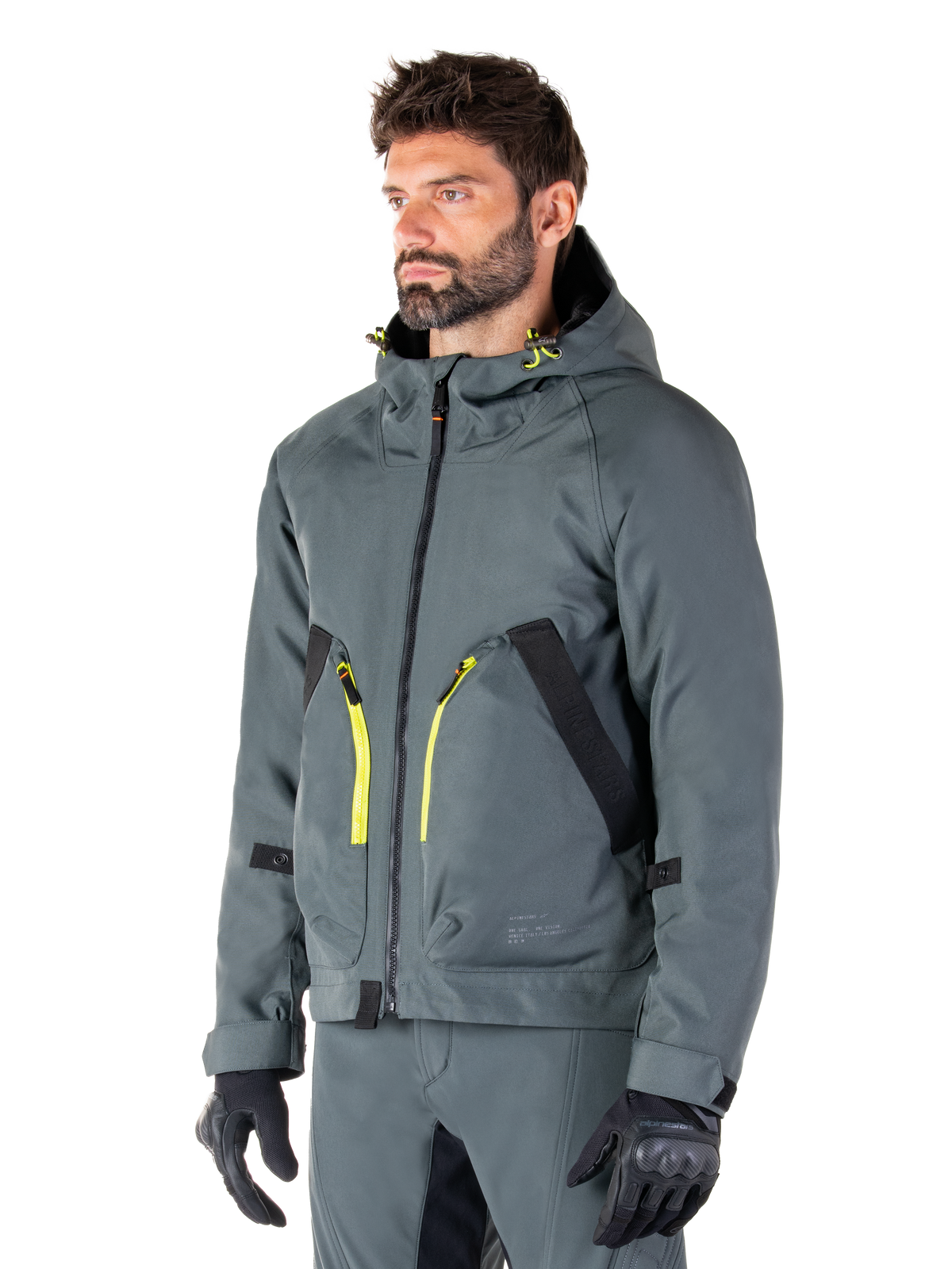 Parka imperméable Alpinestars Morush, veste de moto Urban Gray, design à capuche avec accents de fermetures éclair jaune vif, détails de poches noirs, portée avec un pantalon de moto gris assorti et des gants noirs, vêtement de moto contemporain inspiré du streetwear.