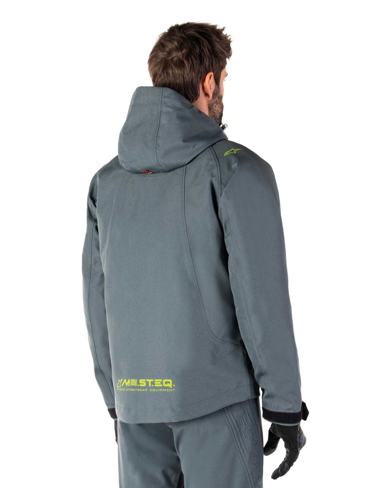 Parka imperméable Alpinestars Morush en Urban Gray, vue de dos, avec marquage MO.ST.EQ. jaune sur le bas du dos et l'épaule, design minimaliste à capuche, portée avec un pantalon de moto gris assorti et des gants noirs, style streetwear moto urbain