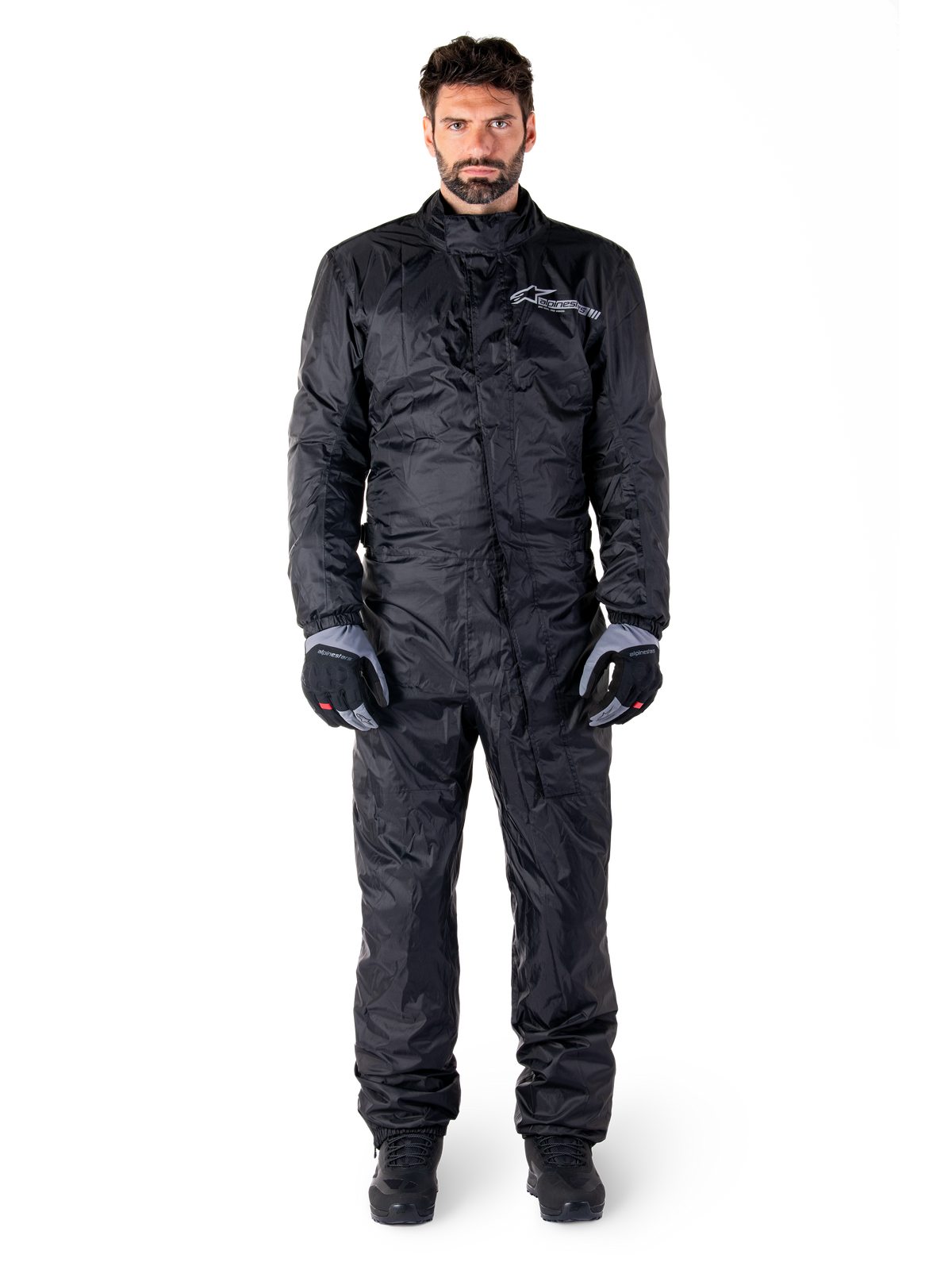 Alpinestars Hurricane Rain V2 Suit, combinaison de pluie intégrale pour moto en noir, équipement de protection imperméable avec col haut et fermeture à rabat-tempête, dotée d'un logo blanc sur la poitrine, portée avec des gants et des bottes noirs et gris