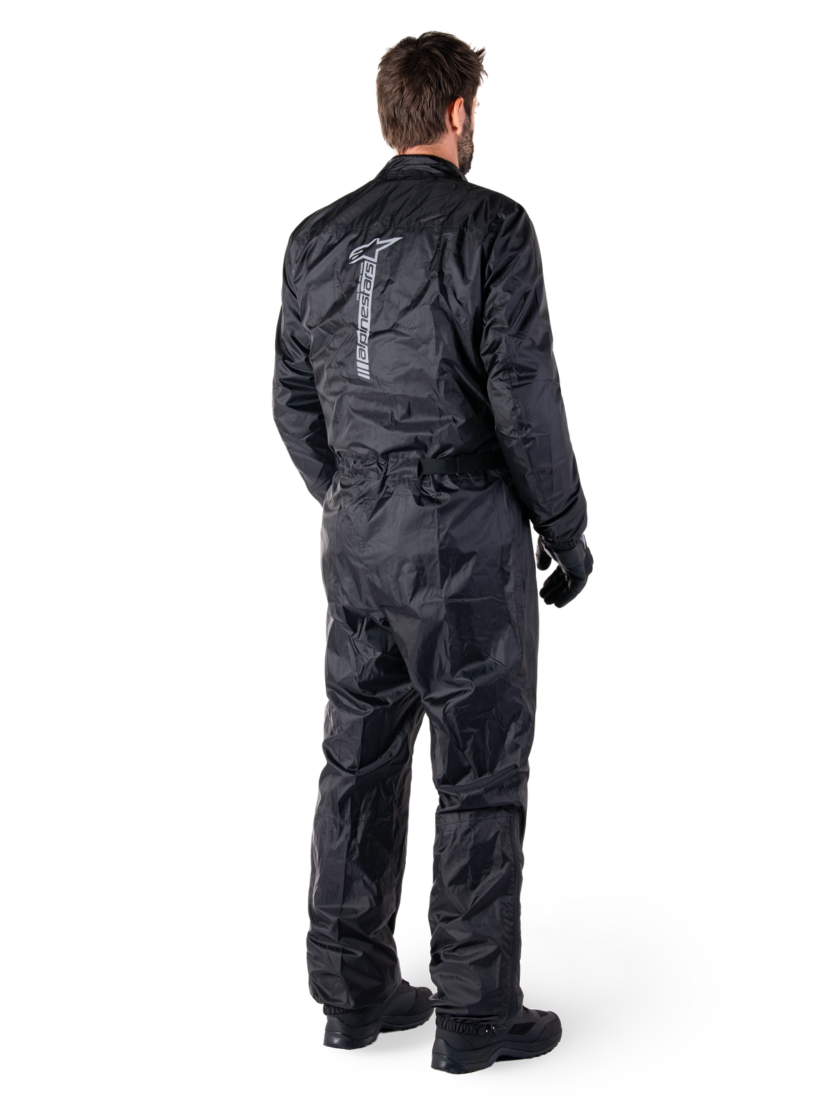 Alpinestars Hurricane Rain V2 Suit, combinaison de pluie moto monopièce imperméable, noire, vue arrière avec grand logo réfléchissant vertical, tissu léger et coupe-vent, portée avec gants et bottes noirs