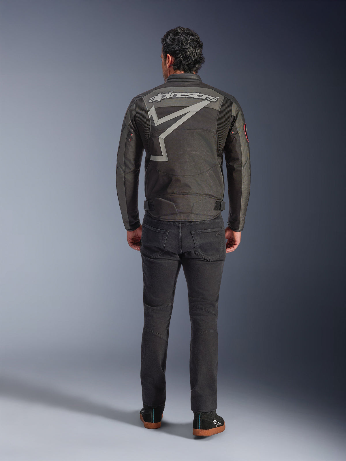 Veste Alpinestars Axton, veste de moto, Noir Anthracite Rouge Tangerine, vue arrière montrant un grand logo Alpinestars gris et un graphisme d'étoile, panneaux accordéon aux épaules, portée avec un pantalon noir et des chaussures noires décontractées.