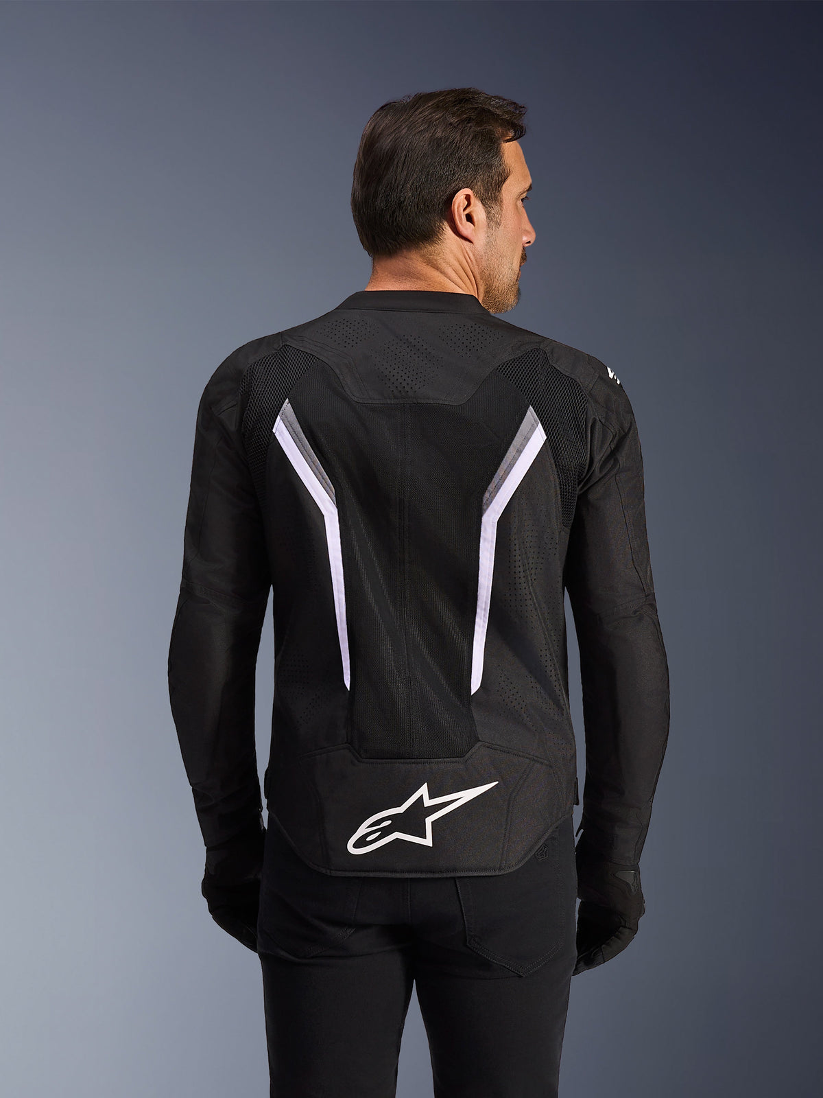 Alpinestars AST V3 Air Jacket, veste de moto, noire avec des accents blancs et gris foncé, vue de dos montrant des panneaux de ventilation en maille étendus, de larges bandes blanches en forme de V et le logo Alpinestars blanc dans le bas du dos, portée avec des gants et un pantalon noirs