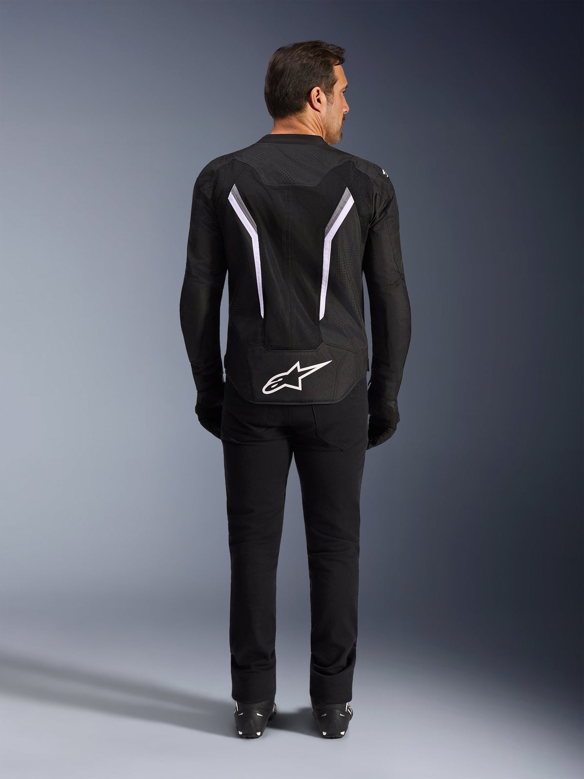 Veste Alpinestars AST V3 Air, vue de dos, noir et gris foncé avec des accents blancs, présentant un grand logo Astar blanc sur le bas du dos, des bandes réfléchissantes et une construction en textile mesh, portée avec un pantalon et des bottes noirs pour un look de conduite sportive