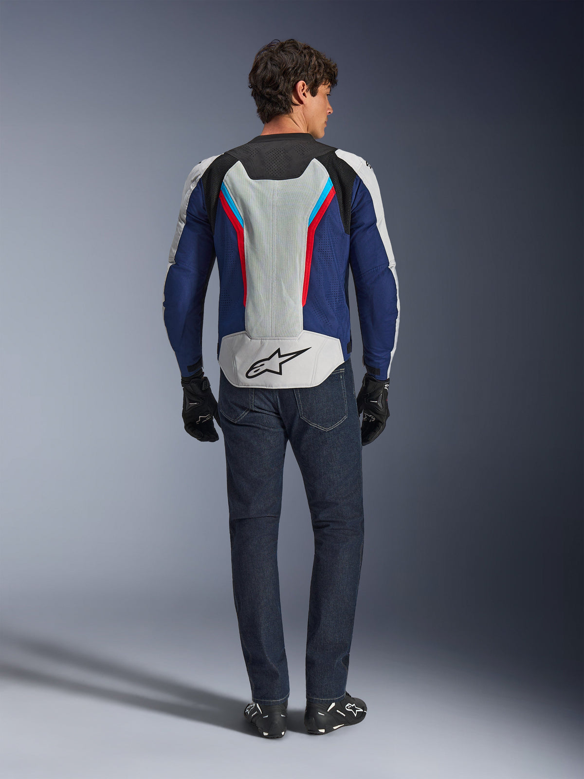 Veste Alpinestars AST V3 Air, vue arrière, Silver Deep Blue et Bright Red, dotée d'un large panneau de ventilation en mesh sur le dos avec le logo Alpinestars, portée avec un jean bleu foncé et des gants noirs, équipement de moto coupe sport pour temps chaud