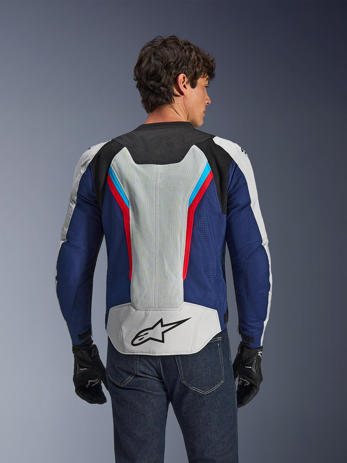 Veste Alpinestars AST V3 Air, veste de moto, argent, bleu profond et rouge éclatant, vue arrière portée par un pilote, dotée de panneaux en maille technique et de perforations laser pour la ventilation, logo étoile noir sur le bas du dos, design sport optimisé pour temps chaud