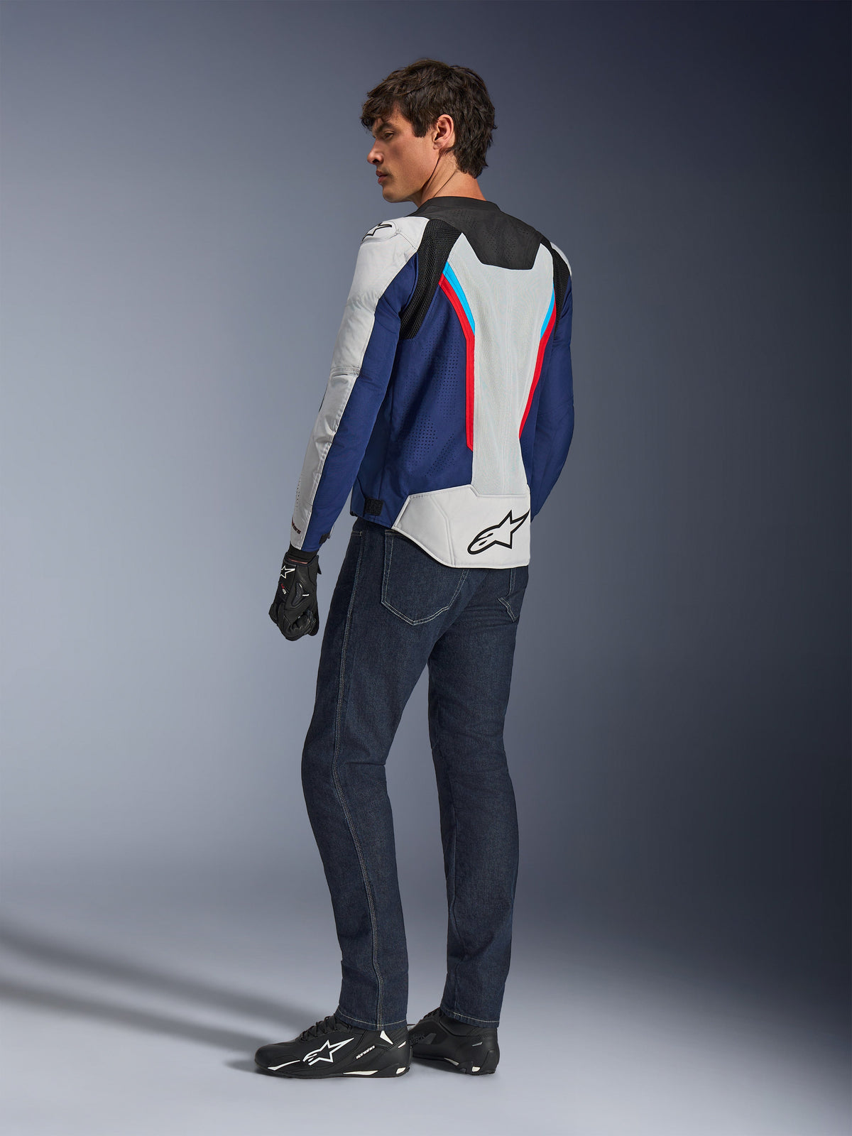 Veste Alpinestars AST V3 Air, veste de moto argentée, bleu foncé et rouge vif, vue de dos portée avec un jean de moto bleu foncé et un équipement noir, dotée de panneaux en maille respirante et du logo Alpinestars dans le dos, équipement de conduite technique à coupe sport.