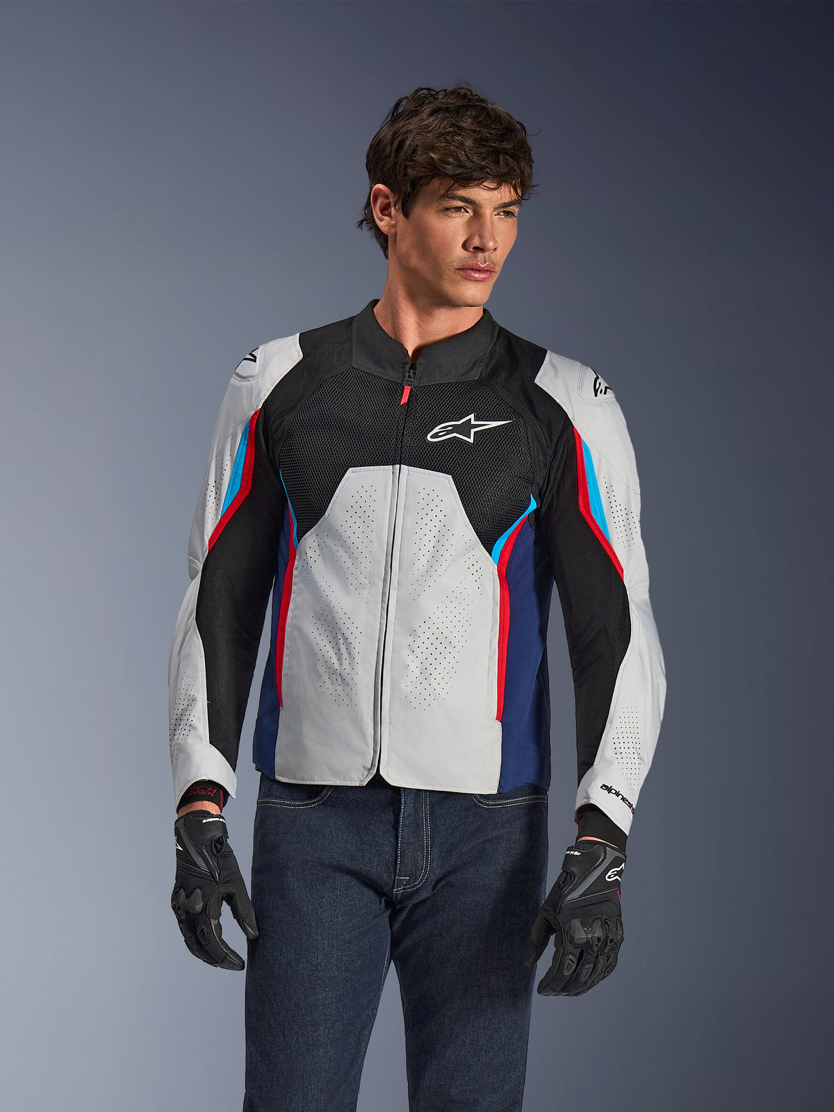 Veste Alpinestars AST V3 Air en Silver Deep Blue Bright Red, portée par un mannequin avec un jean foncé et des gants noirs, dotée de panneaux argentés perforés, de mesh de ventilation noir et d'accents sportifs rouges et bleus.