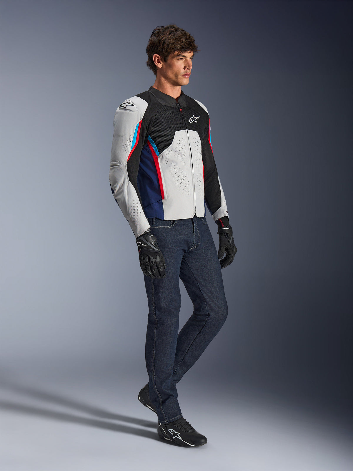 Veste Alpinestars AST V3 Air, veste de moto, argent, bleu foncé et rouge vif, portée avec un jean en denim foncé et des gants de moto noirs, dotée de panneaux en mesh ventilés et d'une coupe sport pour la conduite par temps chaud