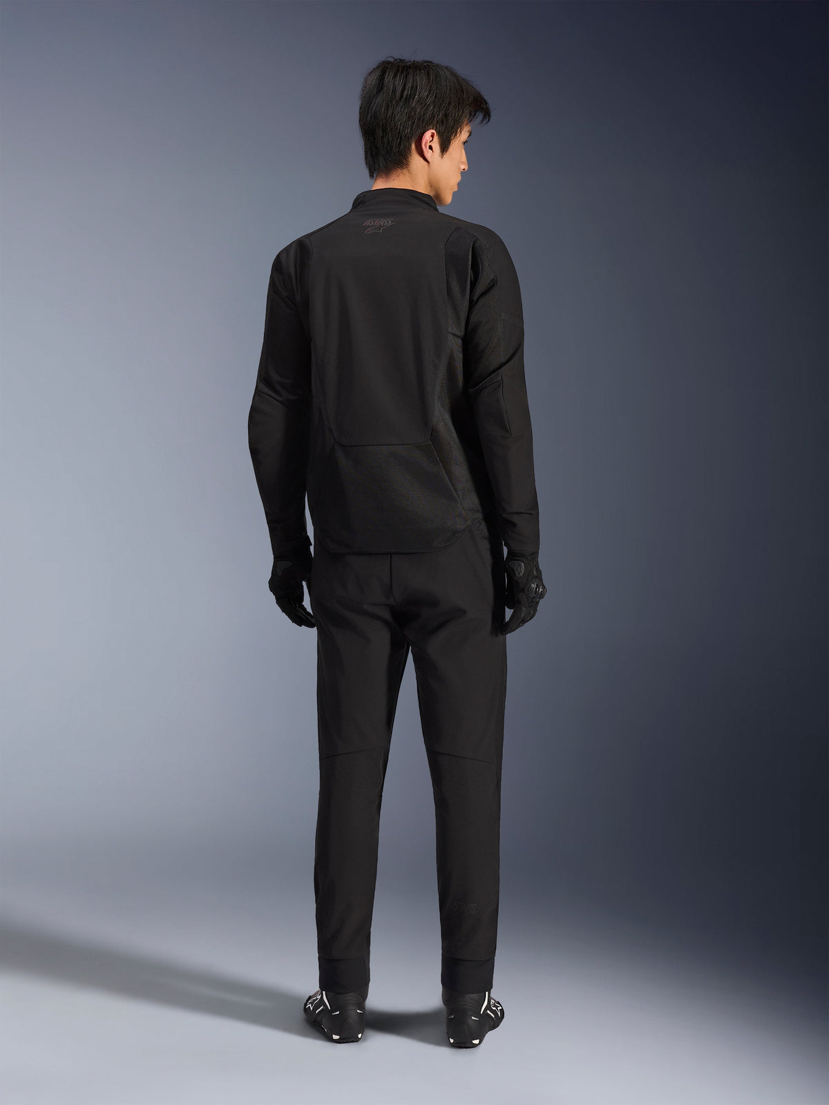 Veste Alpinestars Aeroshell Airflow en Noir Anthracite, vue de dos d'un mannequin homme portant un équipement de moto complet avec pantalon et gants noirs, construction softshell légère avec dos allongé pour une protection accrue.