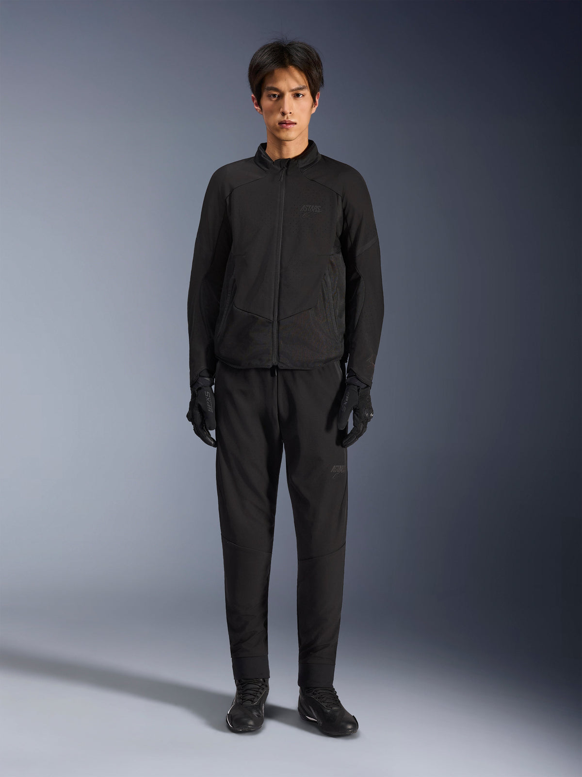 Veste Alpinestars Aeroshell Airflow, veste de moto noire, portée par un mannequin avec un pantalon et des gants noirs assortis, dotée de perforations au laser sur la poitrine et de panneaux latéraux en maille, coupe sport pour une performance par temps chaud