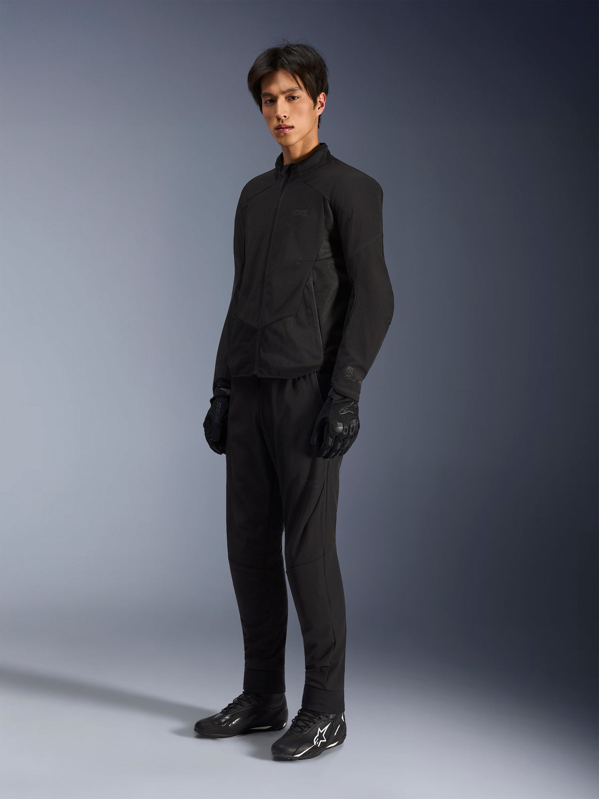 Veste Alpinestars Aeroshell Airflow, veste de moto Black Anthracite, dotée de ventilations perforées au laser et de panneaux en maille pour la circulation de l'air, portée par un mannequin homme avec un pantalon de moto, des gants et des bottes noires assortis avec des logos blancs, coupe sport conçue pour la performance sur route